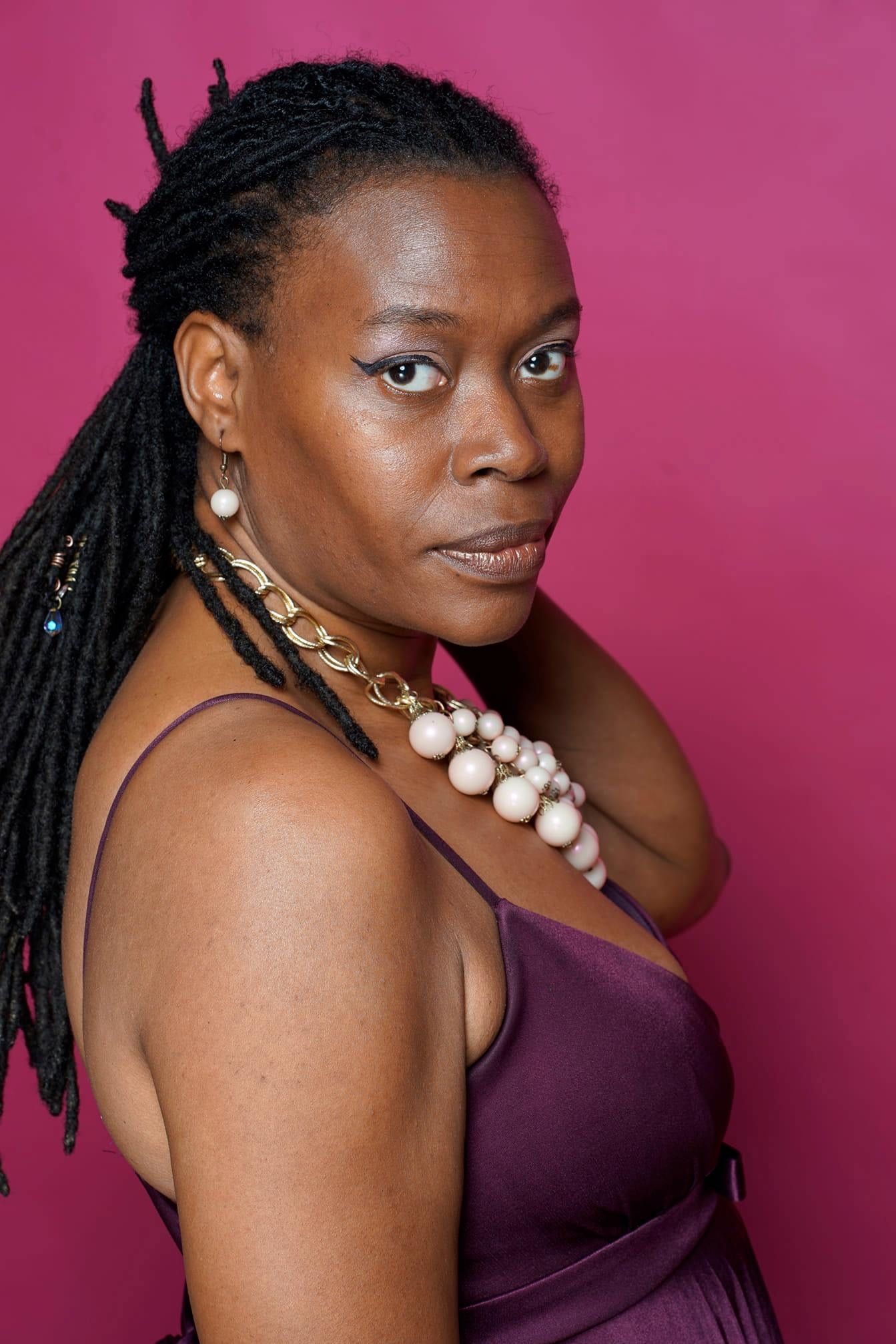 Woman with dreadlocks wearing purple dress and pearl necklace against pink background.