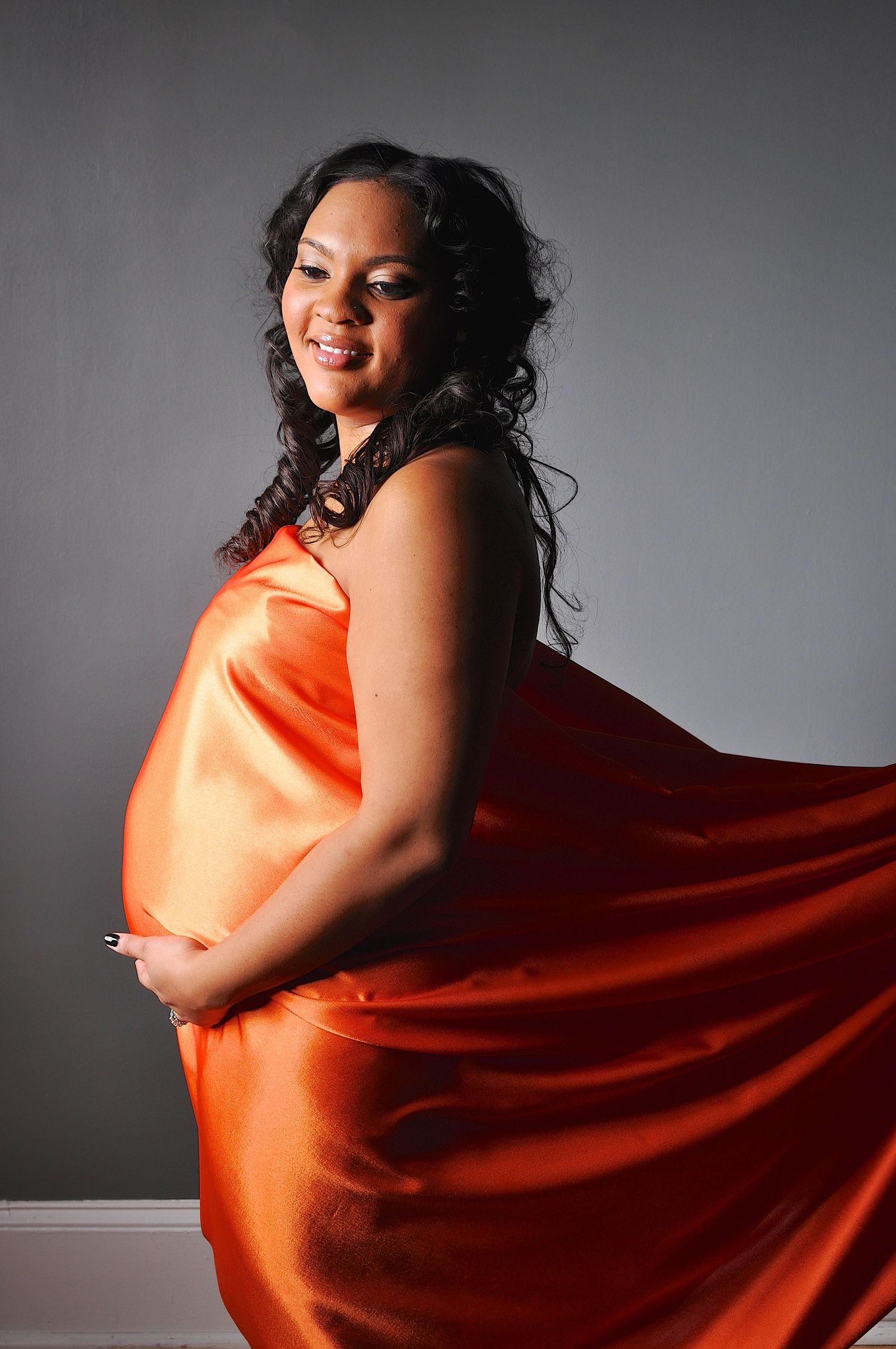 A pregnant woman elegantly draped in flowing orange satin fabric poses against a gray background.