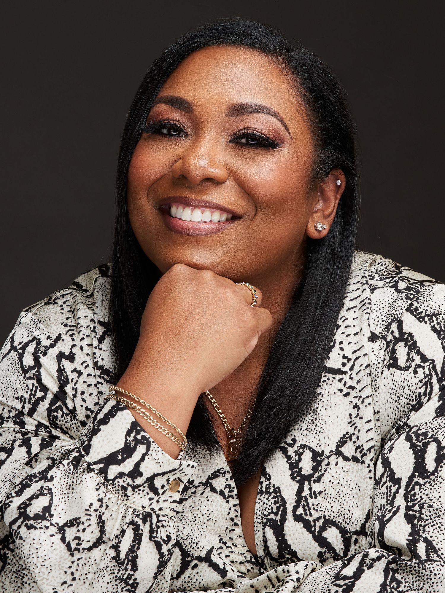 Smiling Black woman in snakeskin blazer with gold jewelry posing against dark background.