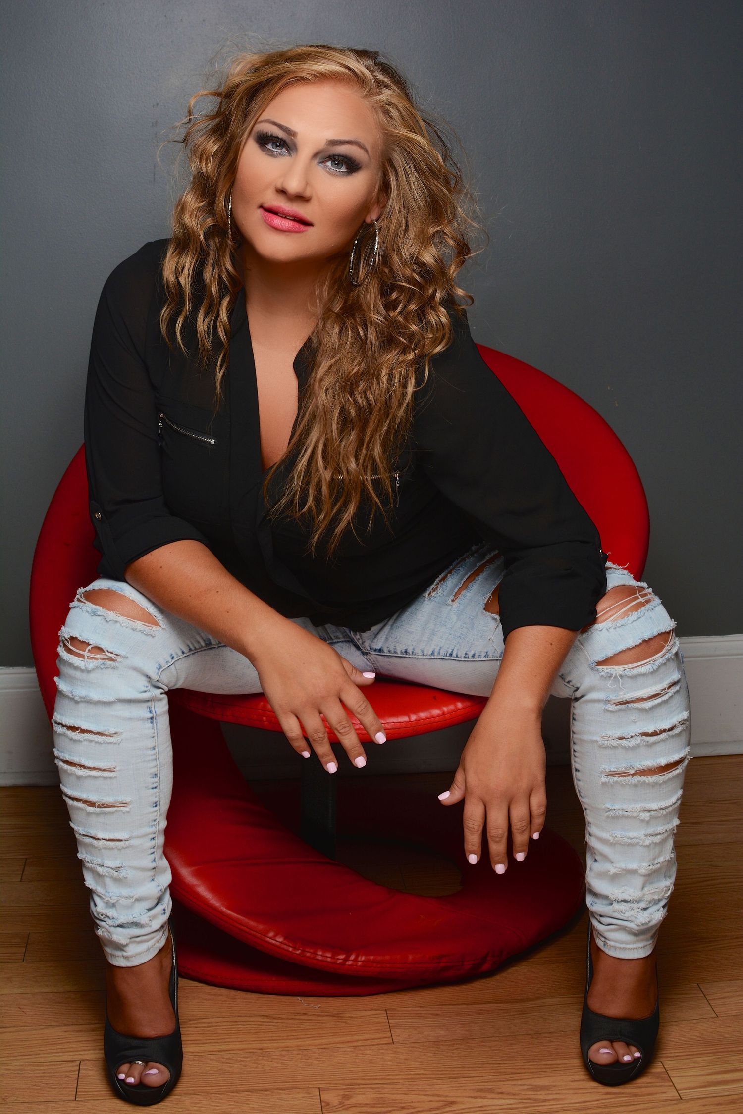 Blonde curly-haired woman in black shirt and ripped jeans sitting on red chair with heels.