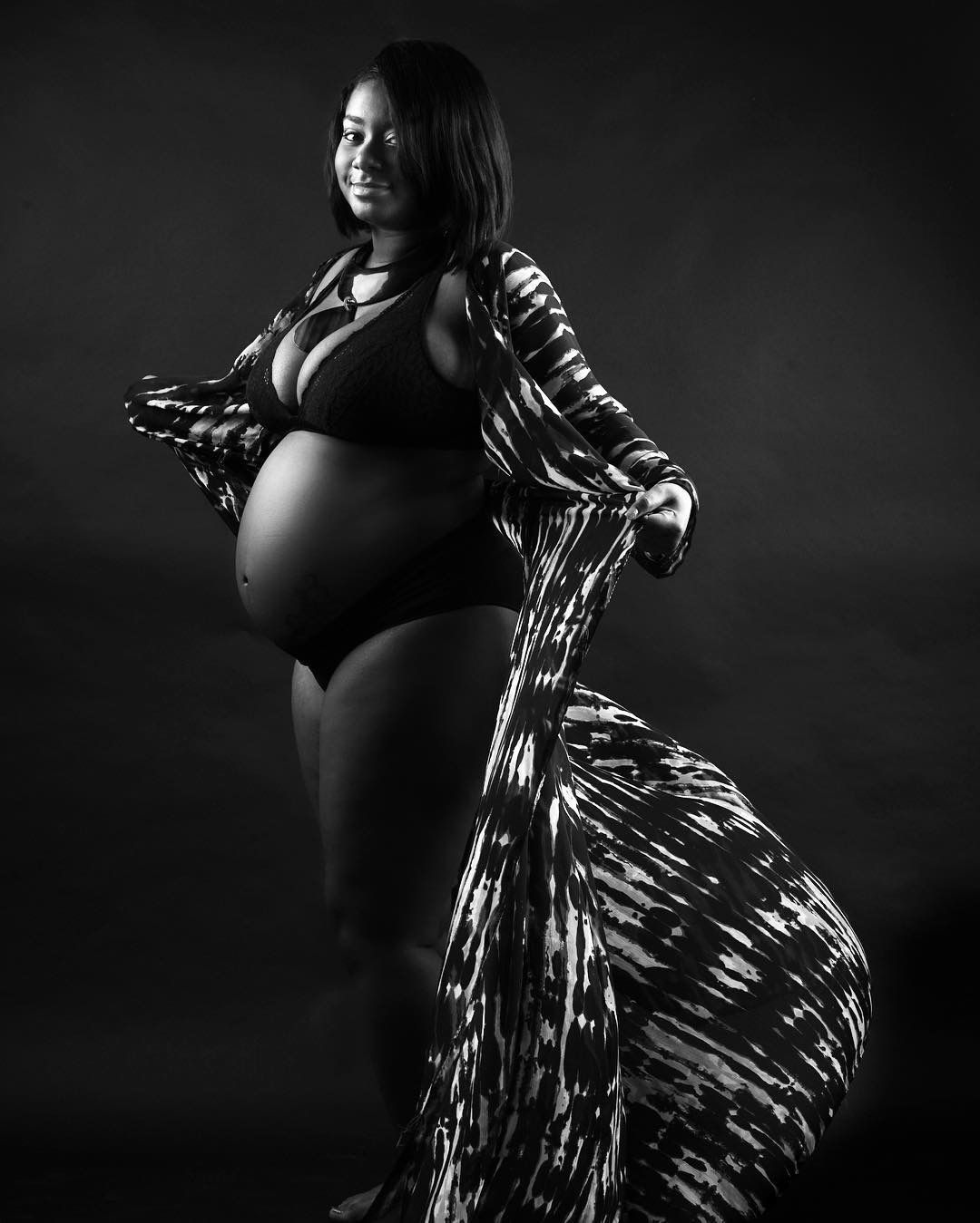 Black and white maternity photo of a pregnant Black woman in a bikini with a flowing patterned robe.