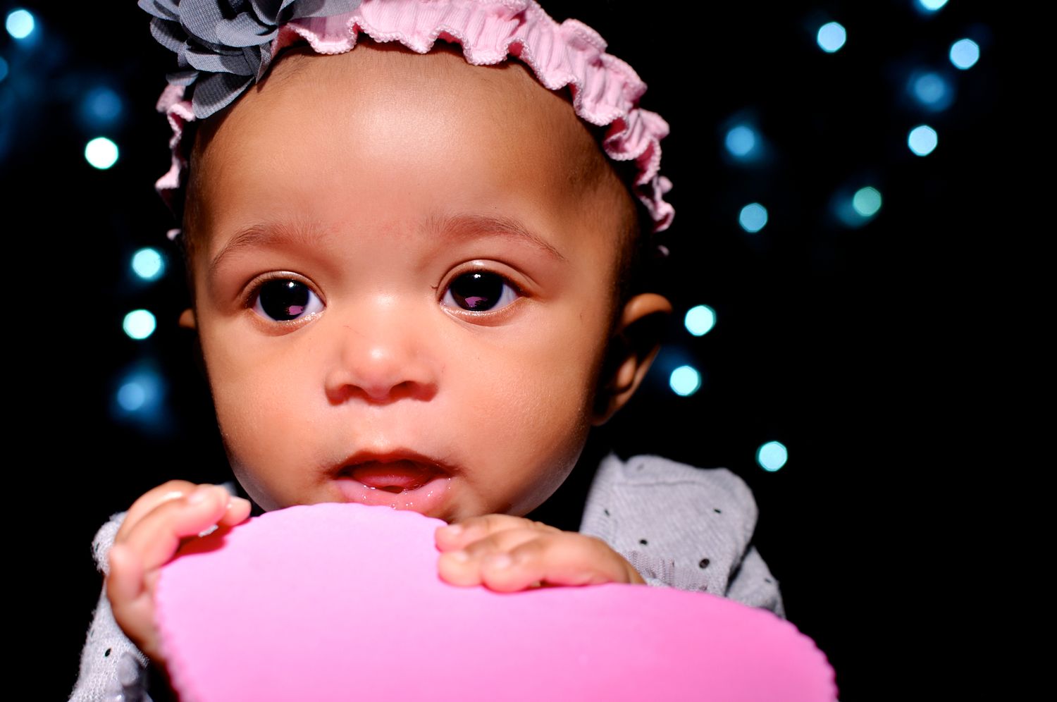 Adorable baby girl with pink bow headband holding a pink heart, with bokeh blue lights in the background.