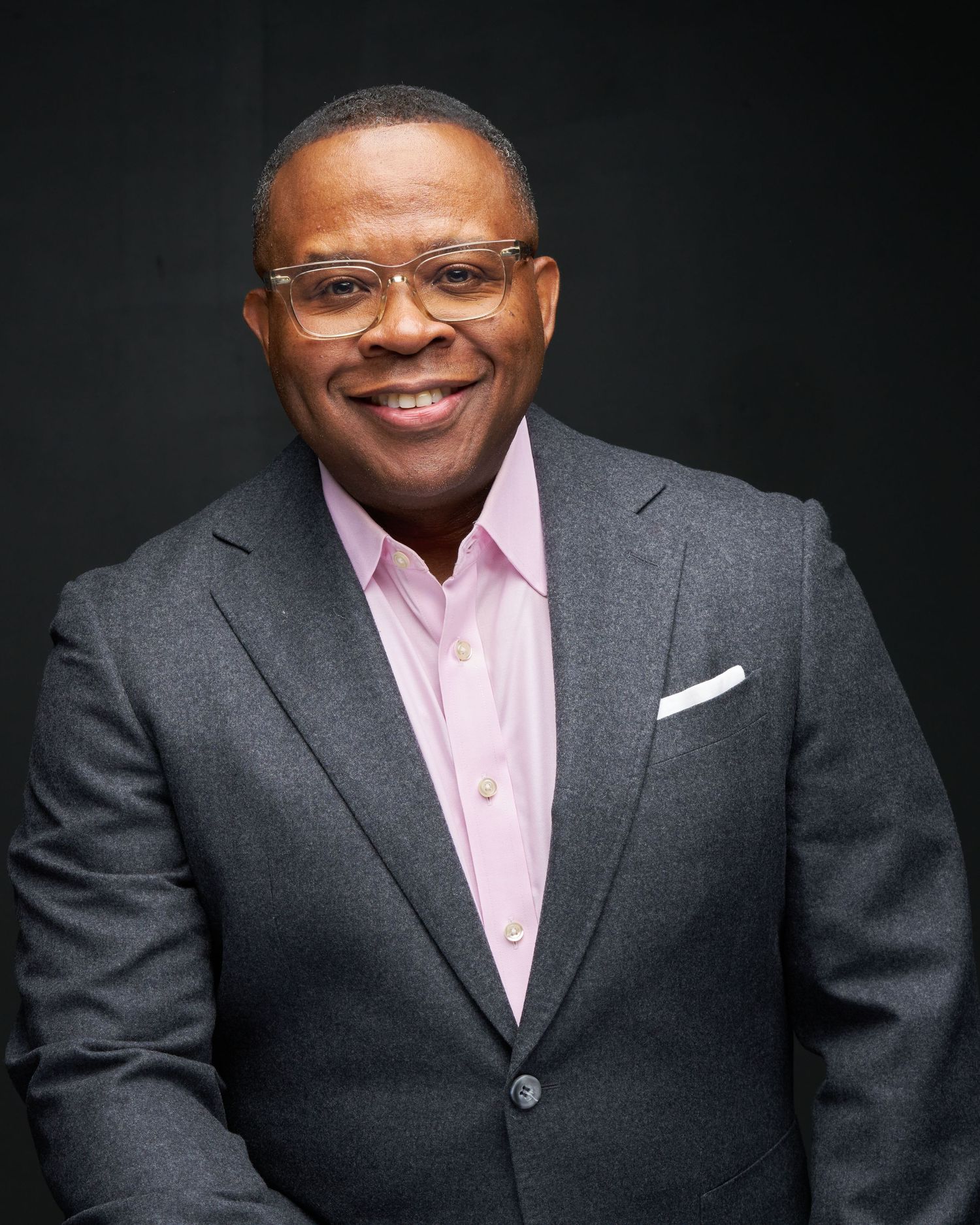 Professional man in gray blazer and pink shirt smiling confidently against dark background.