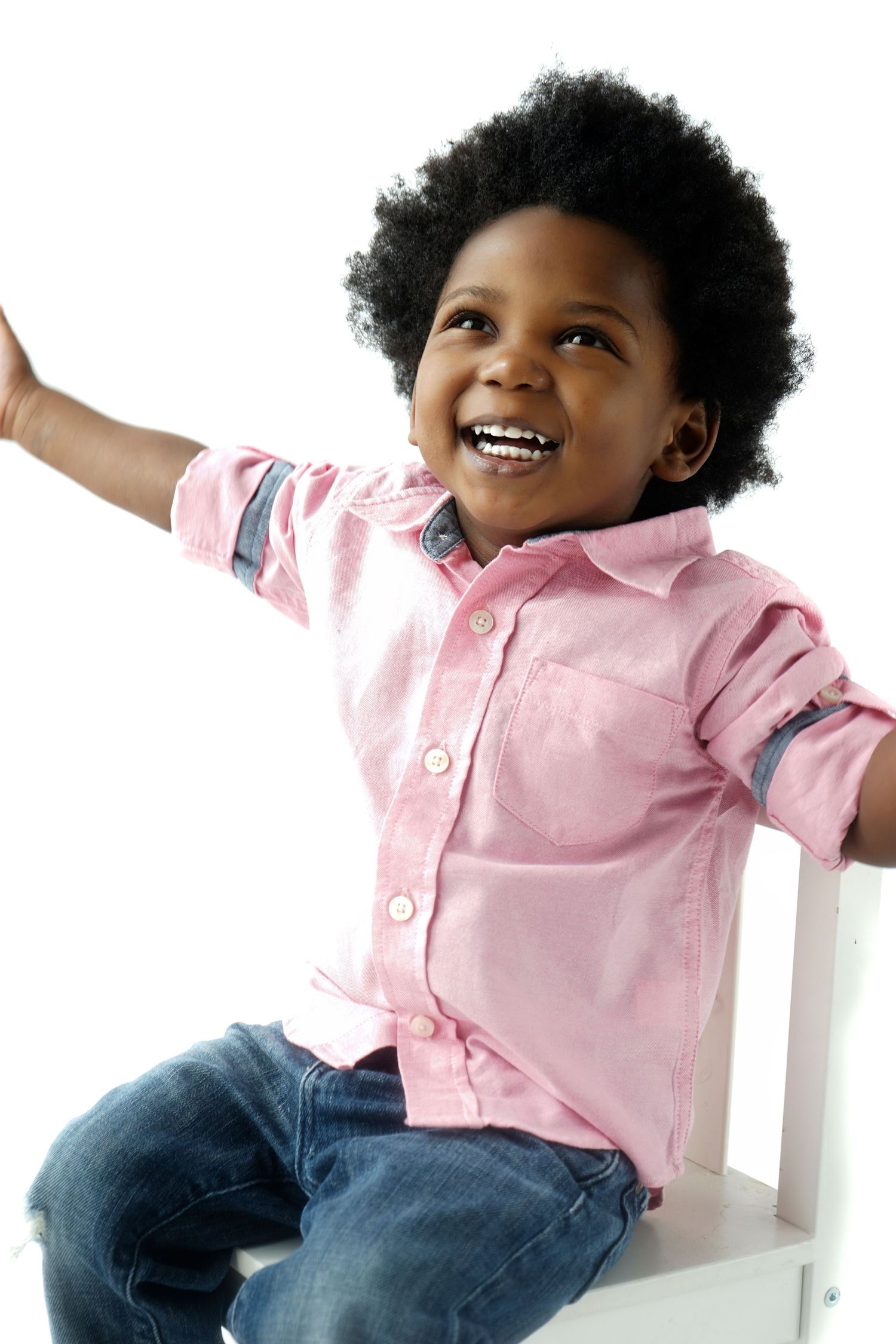 Happy Black toddler boy with afro wearing pink shirt and jeans sitting on white chair laughing.