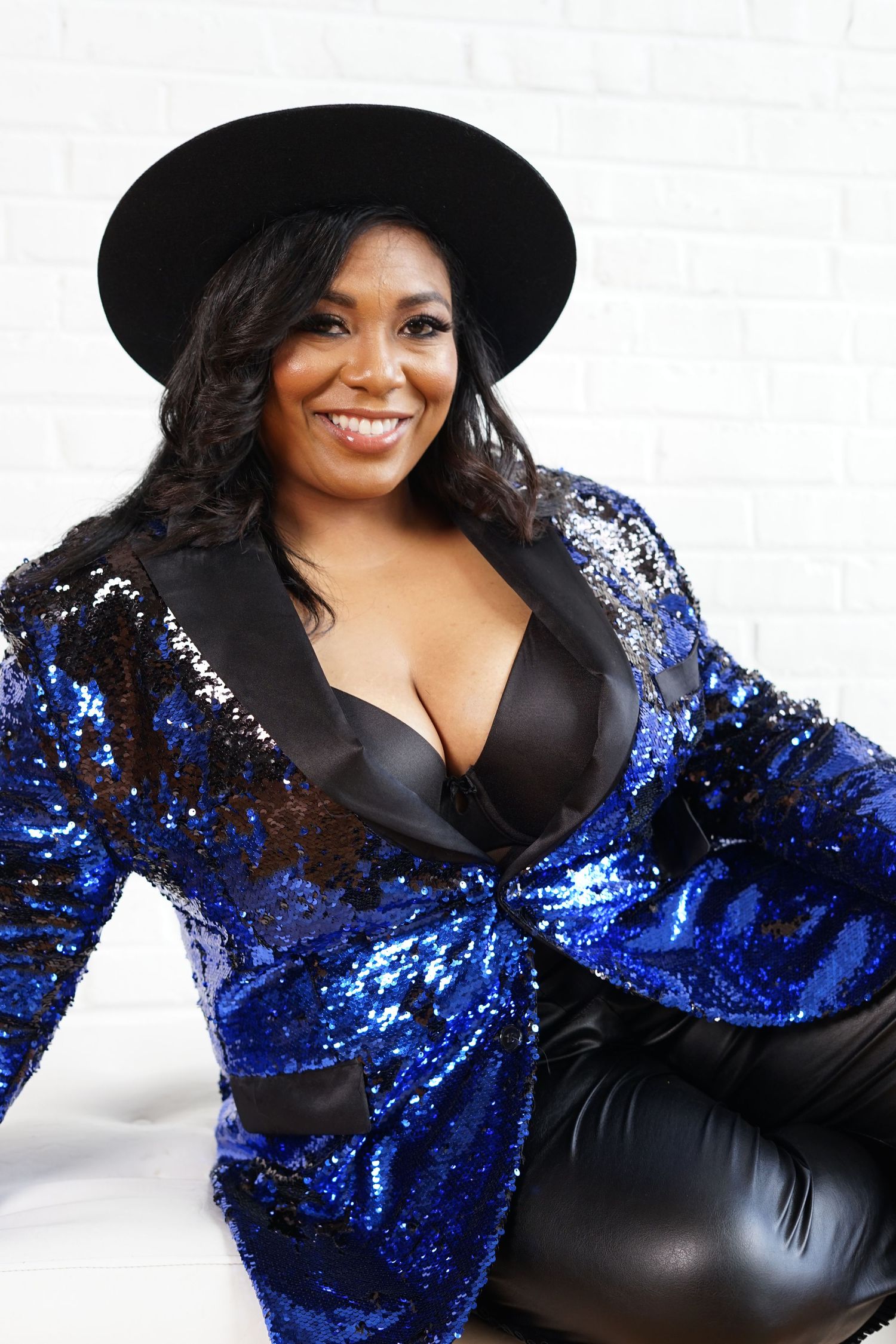 A confident plus-size Black woman poses in a blue sequin blazer and black hat against a white brick wall.