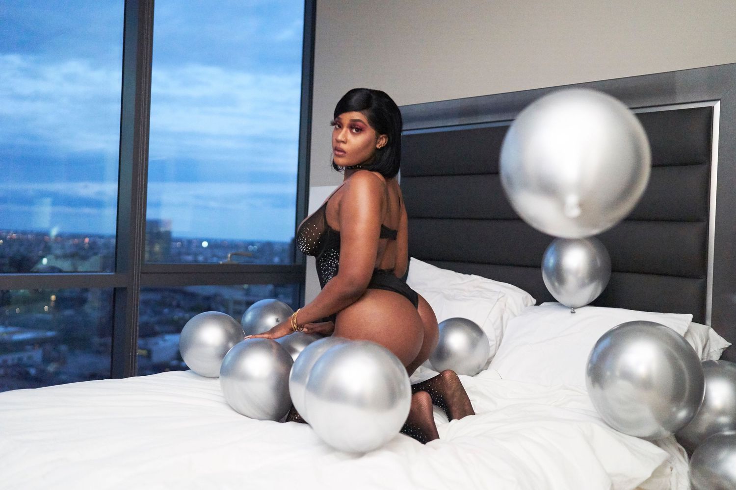 A young Black woman in black lingerie poses on a bed surrounded by silver balloons near a city view window.