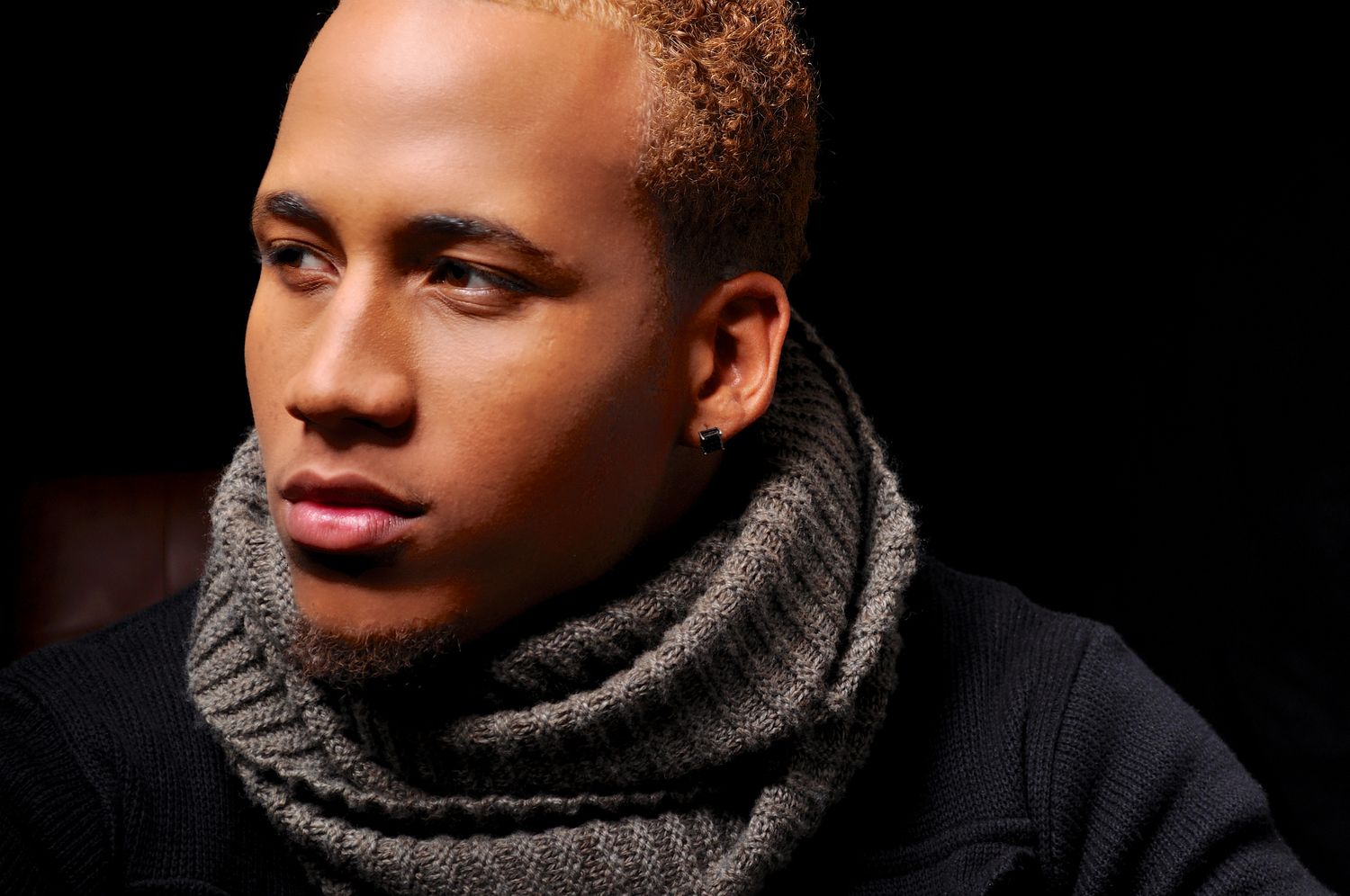 Young mixed-race man with blonde cropped hair wearing gray knit scarf in moody low-light portrait.