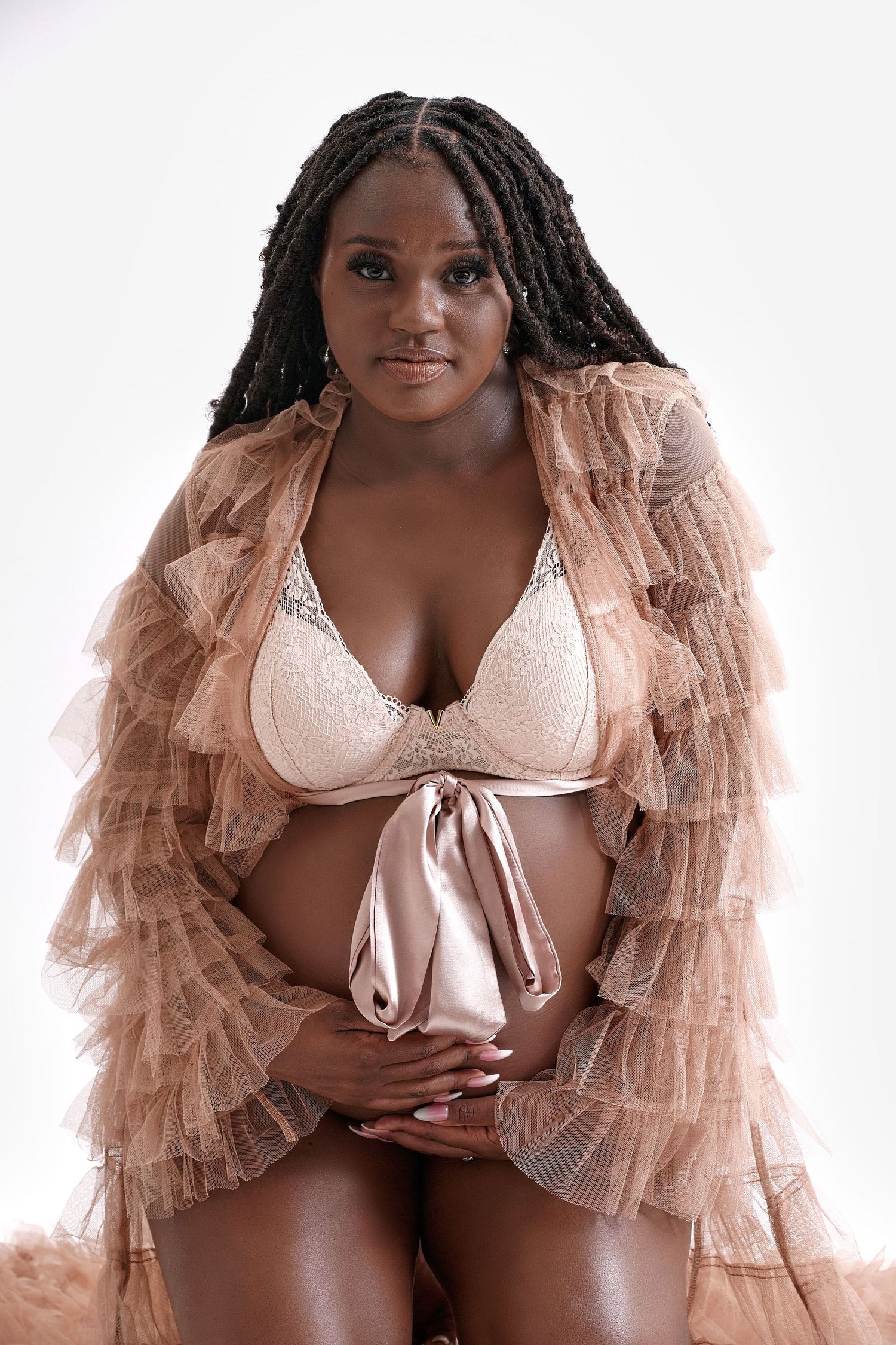 Pregnant woman in beige lace lingerie and ruffled robe cradling baby bump.