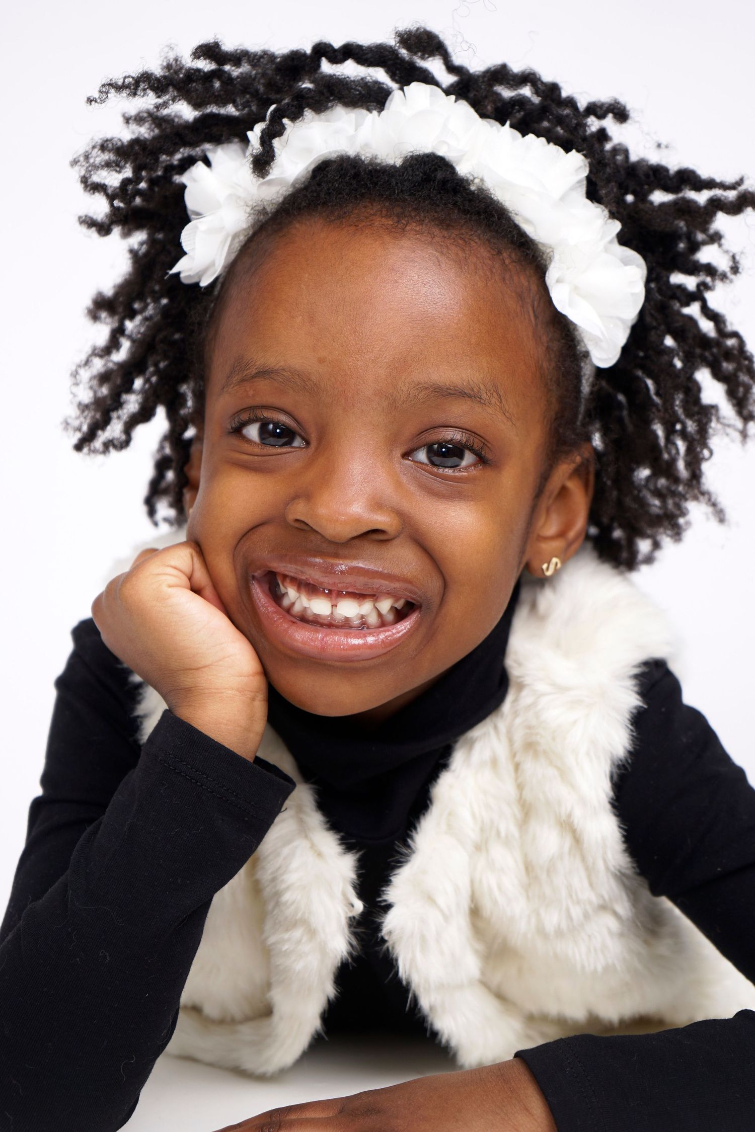 Happy young Black girl with braided pigtails and white bows wearing a black turtleneck and fur vest.