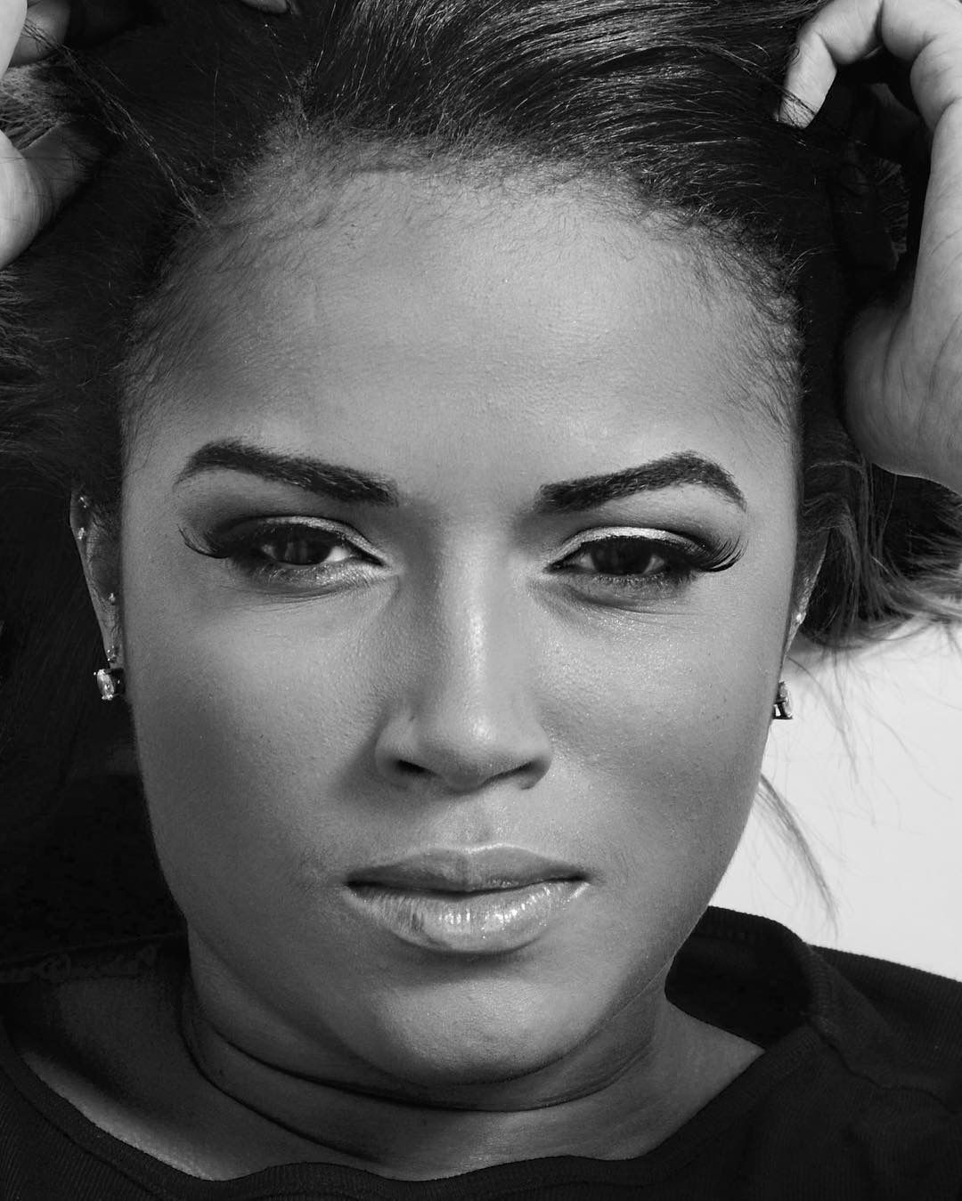 Black and white close-up portrait of a young woman with dramatic eye makeup and hands in hair.