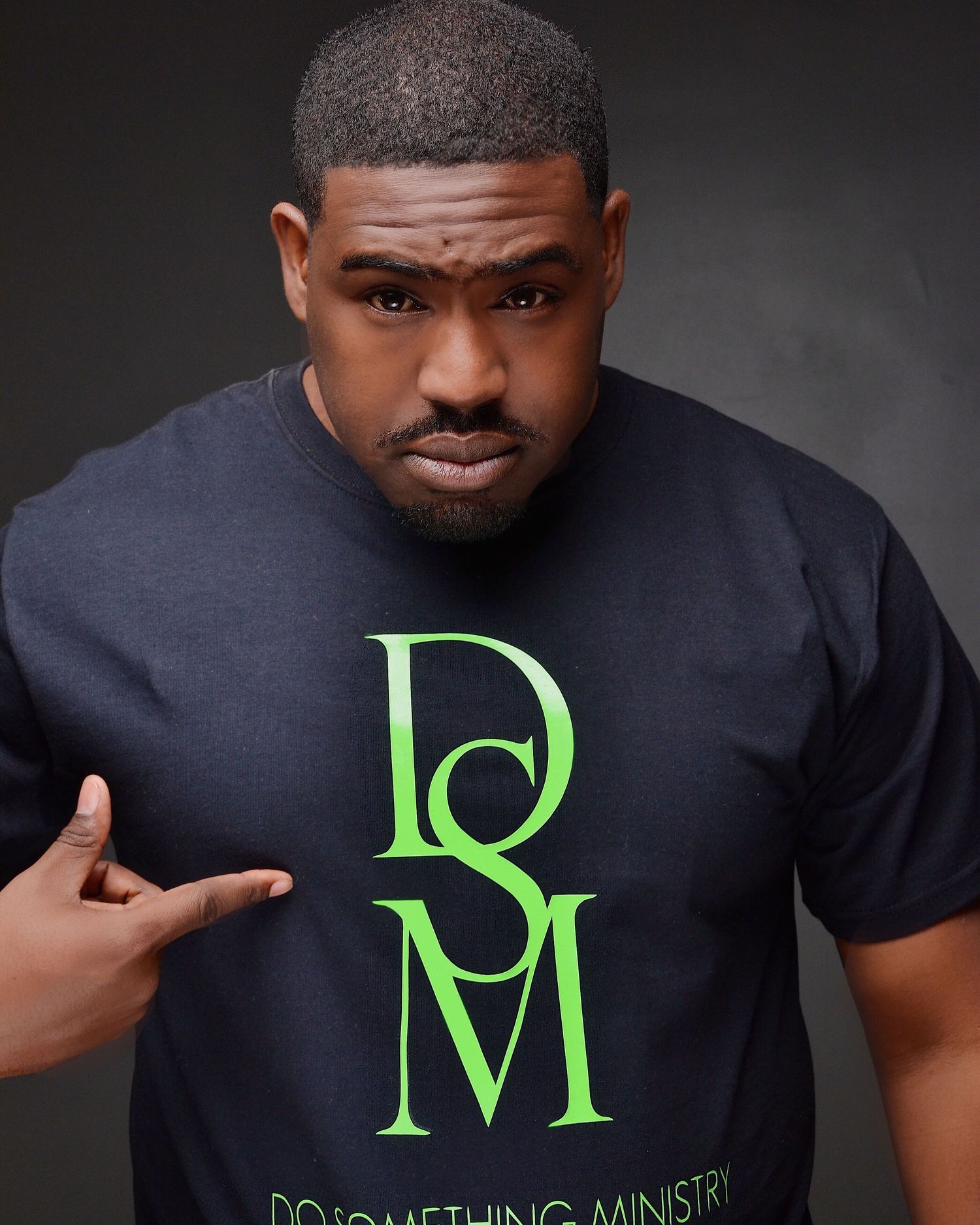 Man in a black DSM Do Something Ministry t-shirt pointing at logo against dark background.