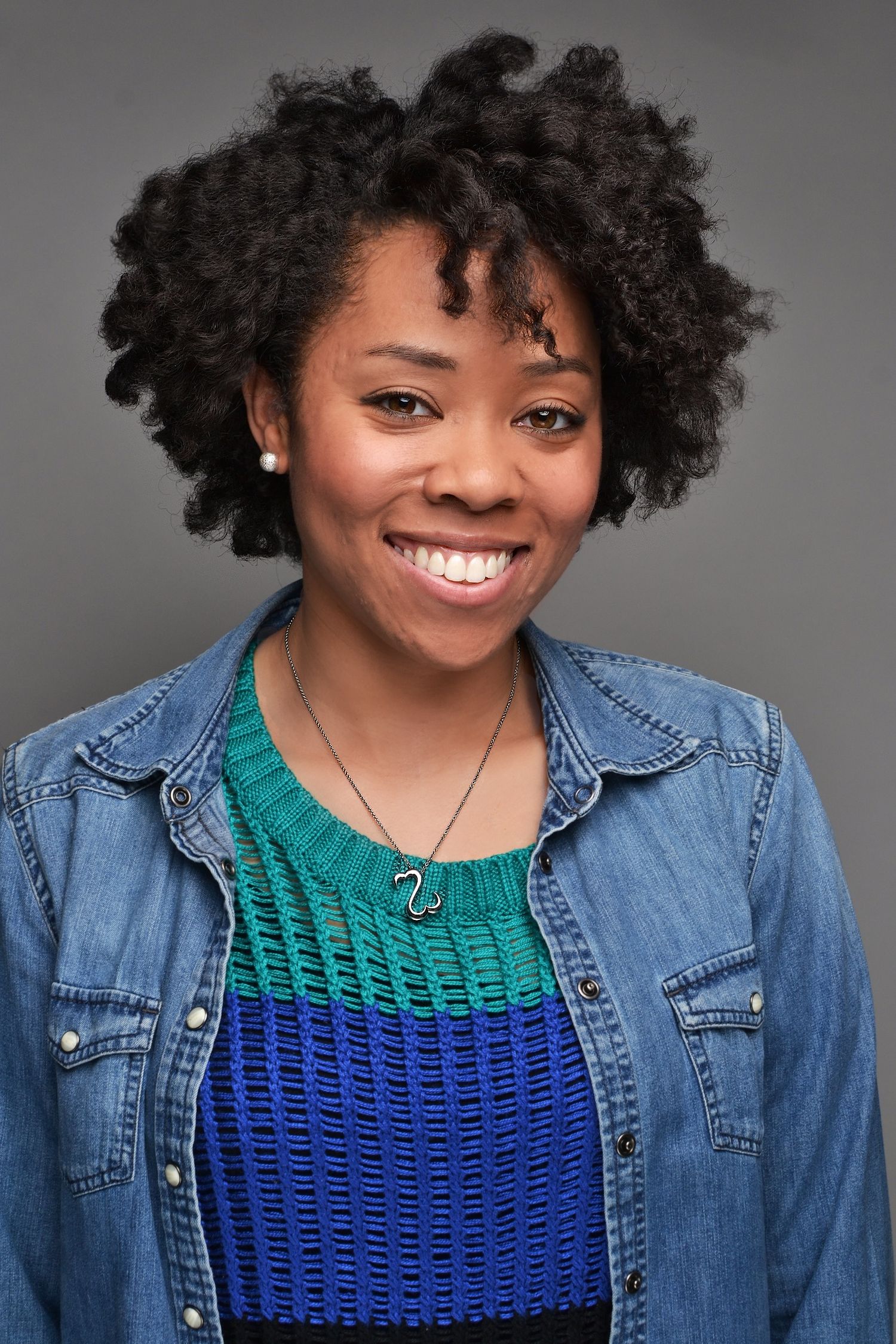 Smiling young Black woman with natural curly hair wearing denim jacket and blue-green sweater.