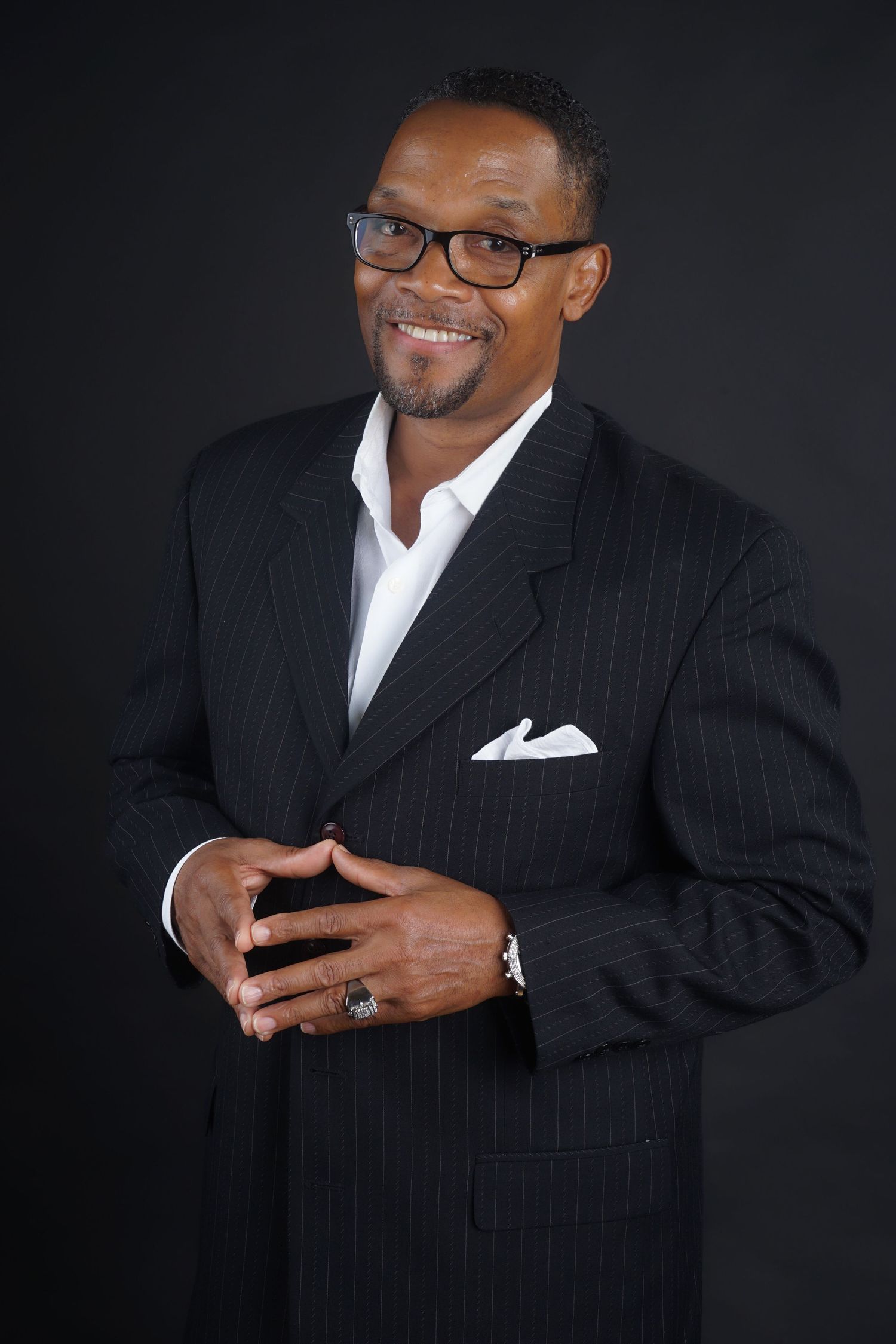 Smiling man in pinstripe suit with white pocket square and glasses poses confidently against dark background.