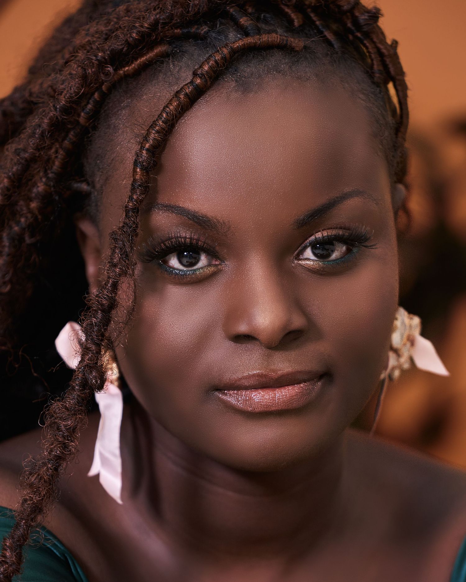 A young woman with braided locs and pink flower earrings poses in a warm-toned portrait photo.