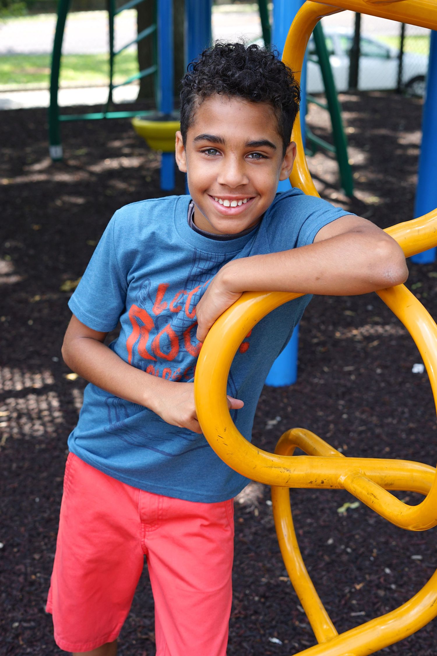 Smiling boy in blue shirt and red shorts leaning on yellow playground equipment.