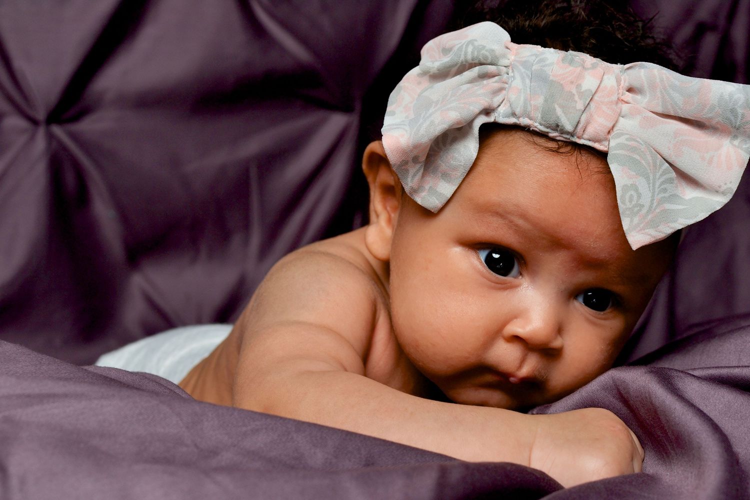 Adorable baby girl wearing a floral bow headband lying on a purple satin background.