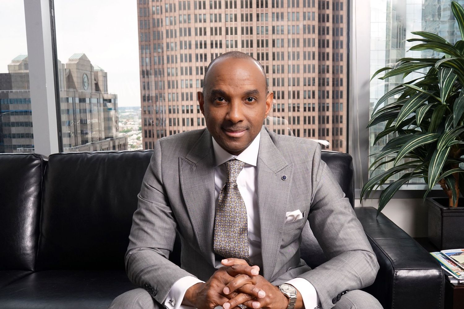 Bald Black man in gray suit sitting on leather sofa with city skyline view in background.
