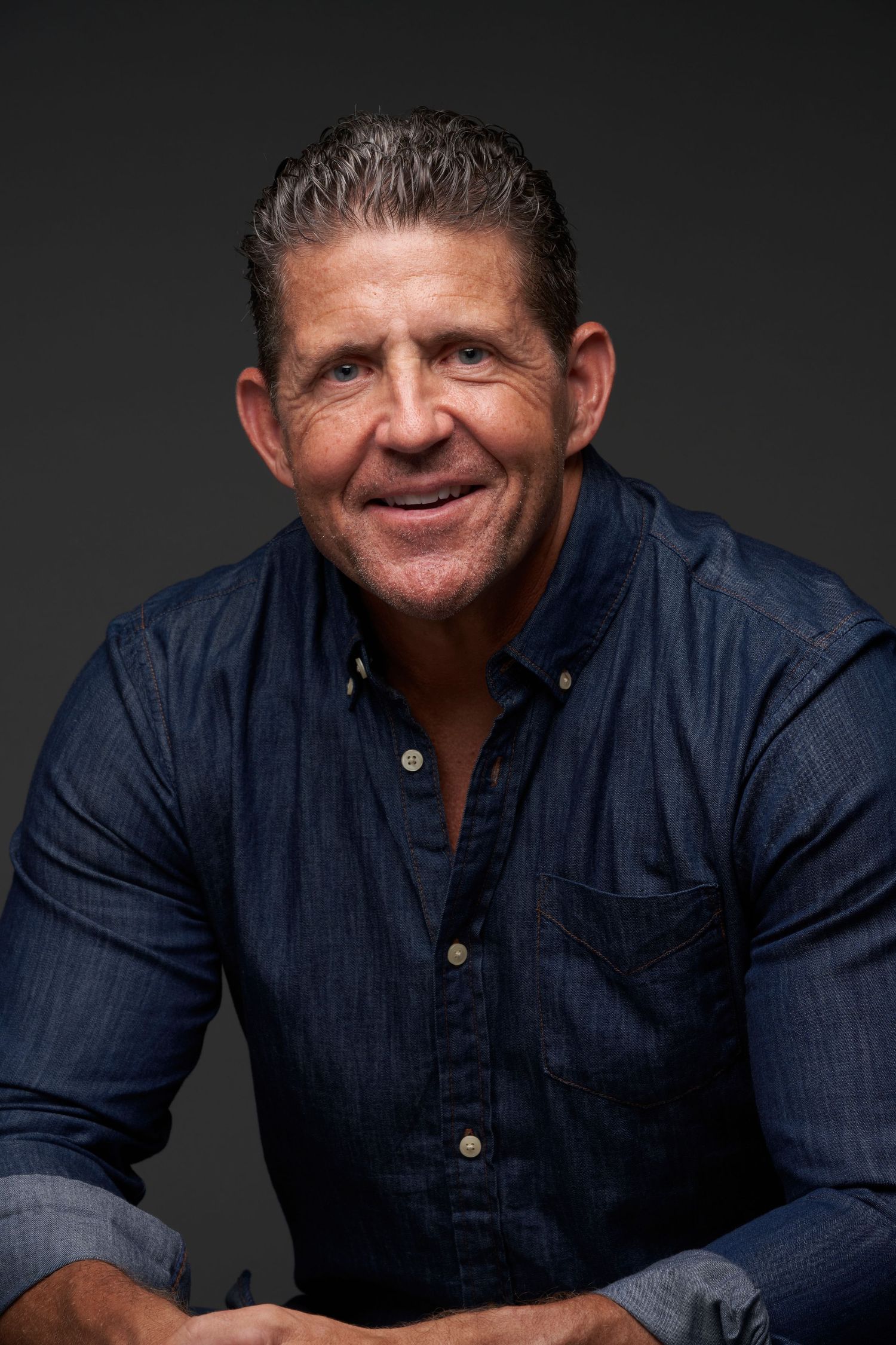 A middle-aged man in a dark denim shirt smiles warmly during a professional studio portrait session.