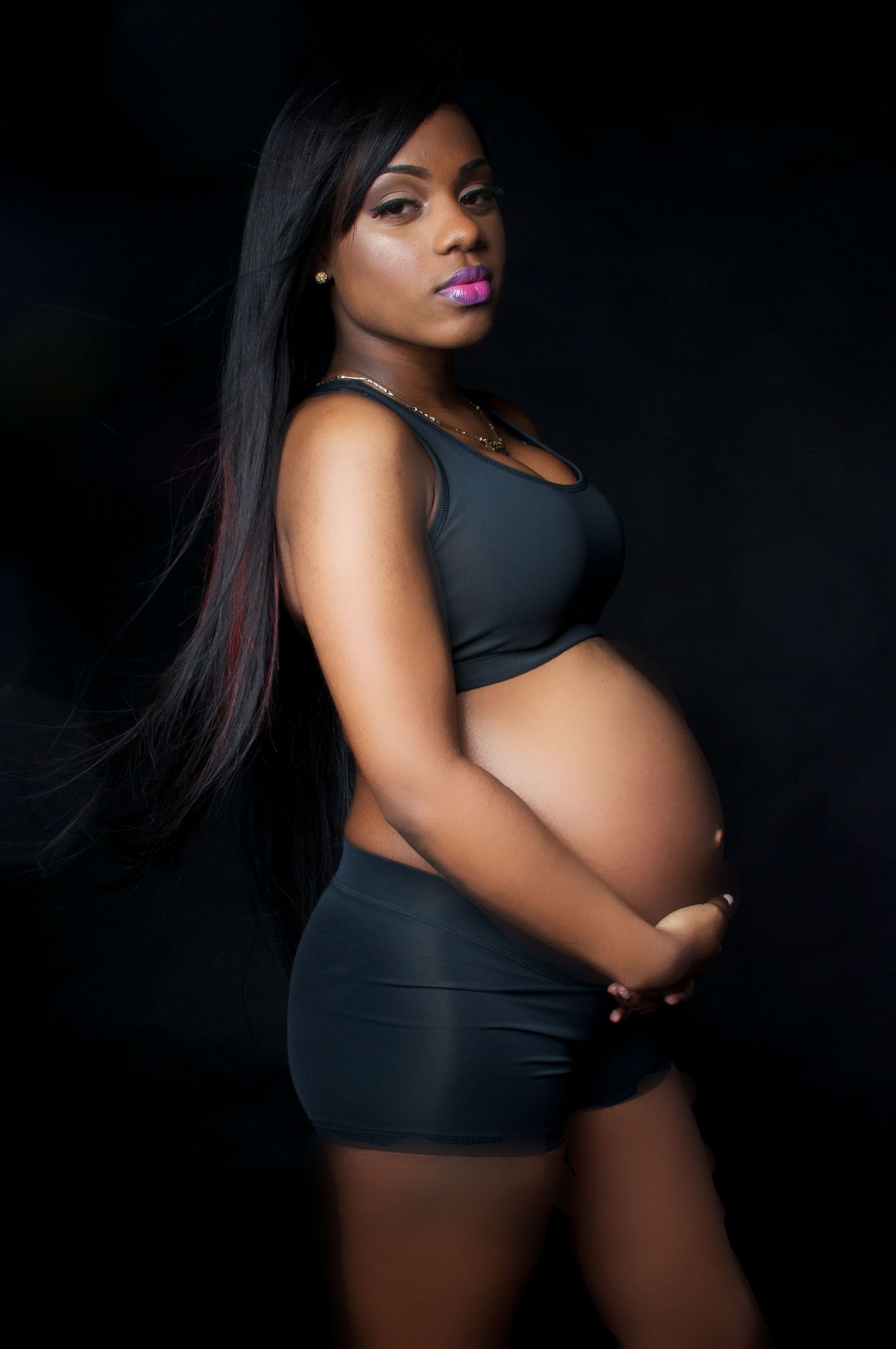 Pregnant woman in a dark outfit holding her baby bump during a dramatic studio portrait session.