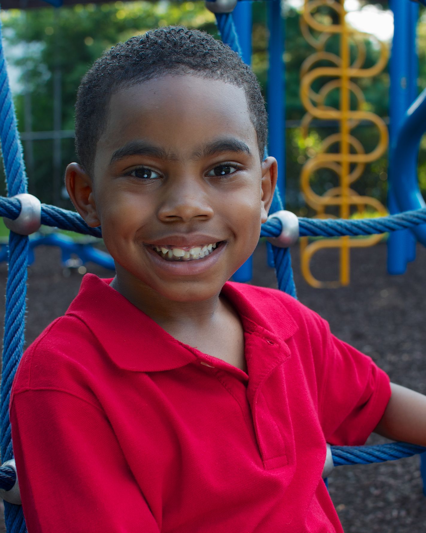 Happy Black boy in a red polo shirt smiling on a colorful playground climbing structure.