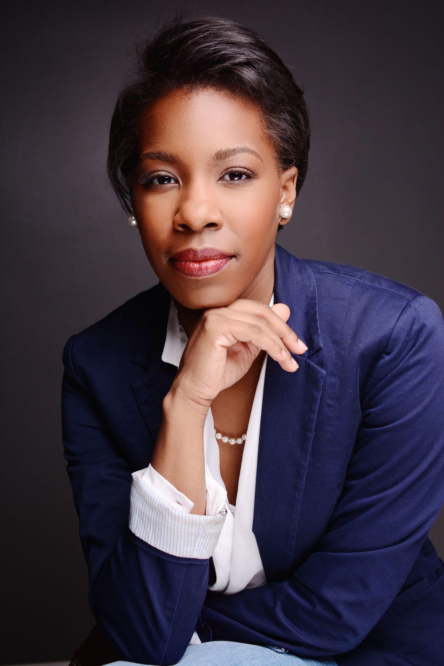 Professional headshot of a woman in a navy blazer and white blouse, posing with chin resting on hands.