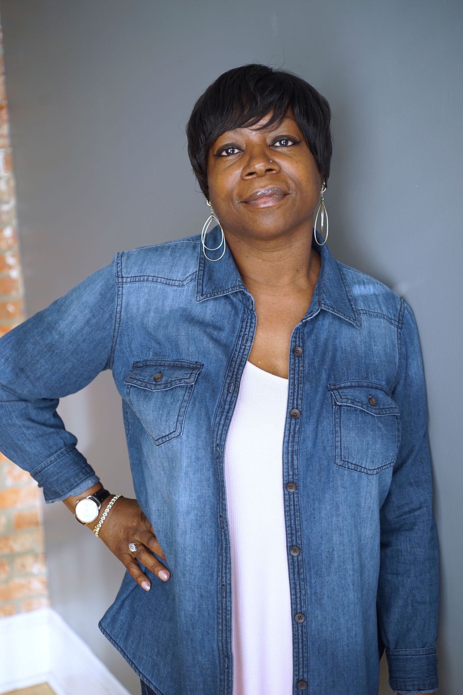 Middle-aged Black woman in denim shirt posing with hand on hip, casual portrait against gray wall.