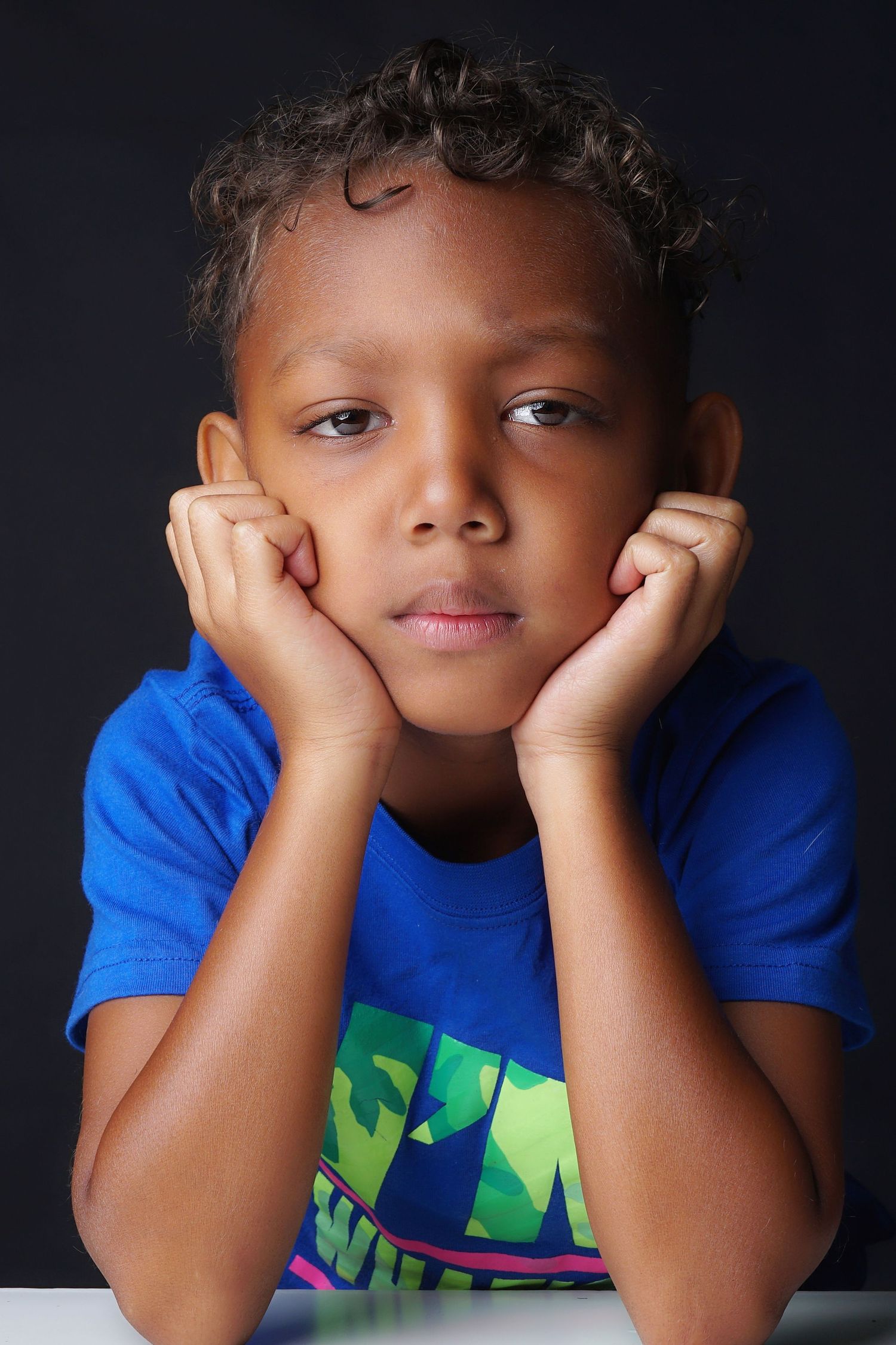Young boy in blue t-shirt resting chin on hands with thoughtful expression.