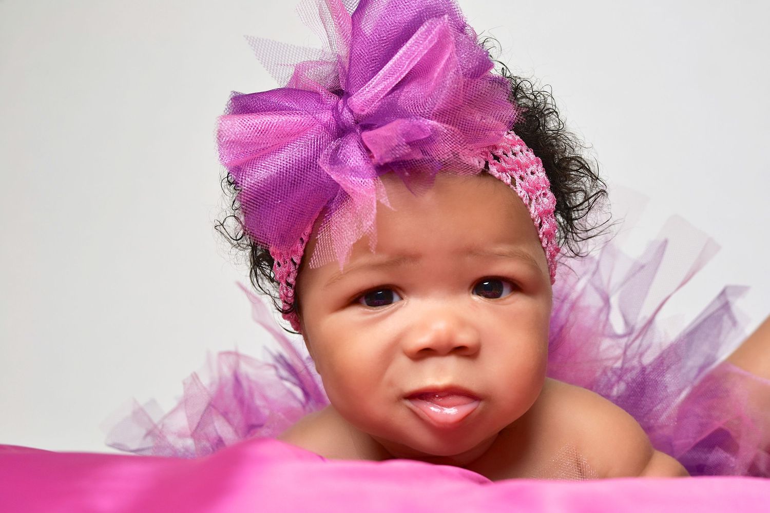 Cute Black baby girl wearing a large purple tutu bow headband lying on pink fabric.