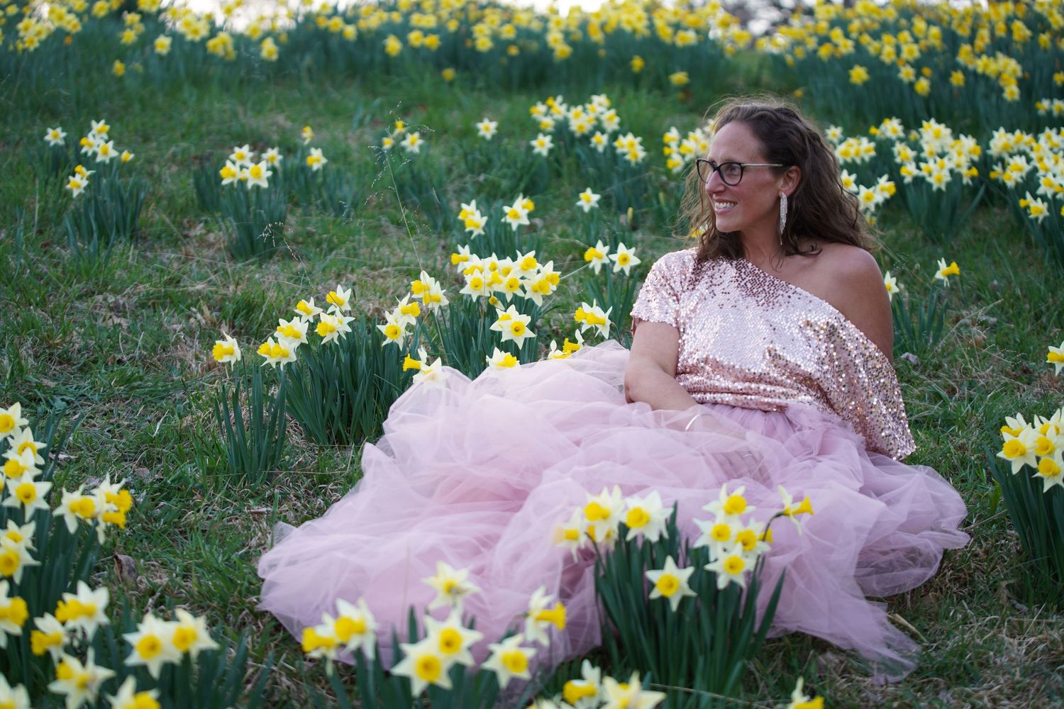 Woman in pink tulle gown sitting among yellow daffodils in outdoor field.