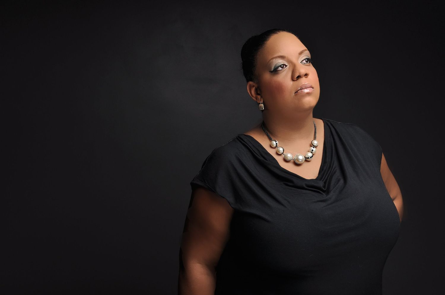 A woman in a black top and pearl necklace poses confidently against a dark background.