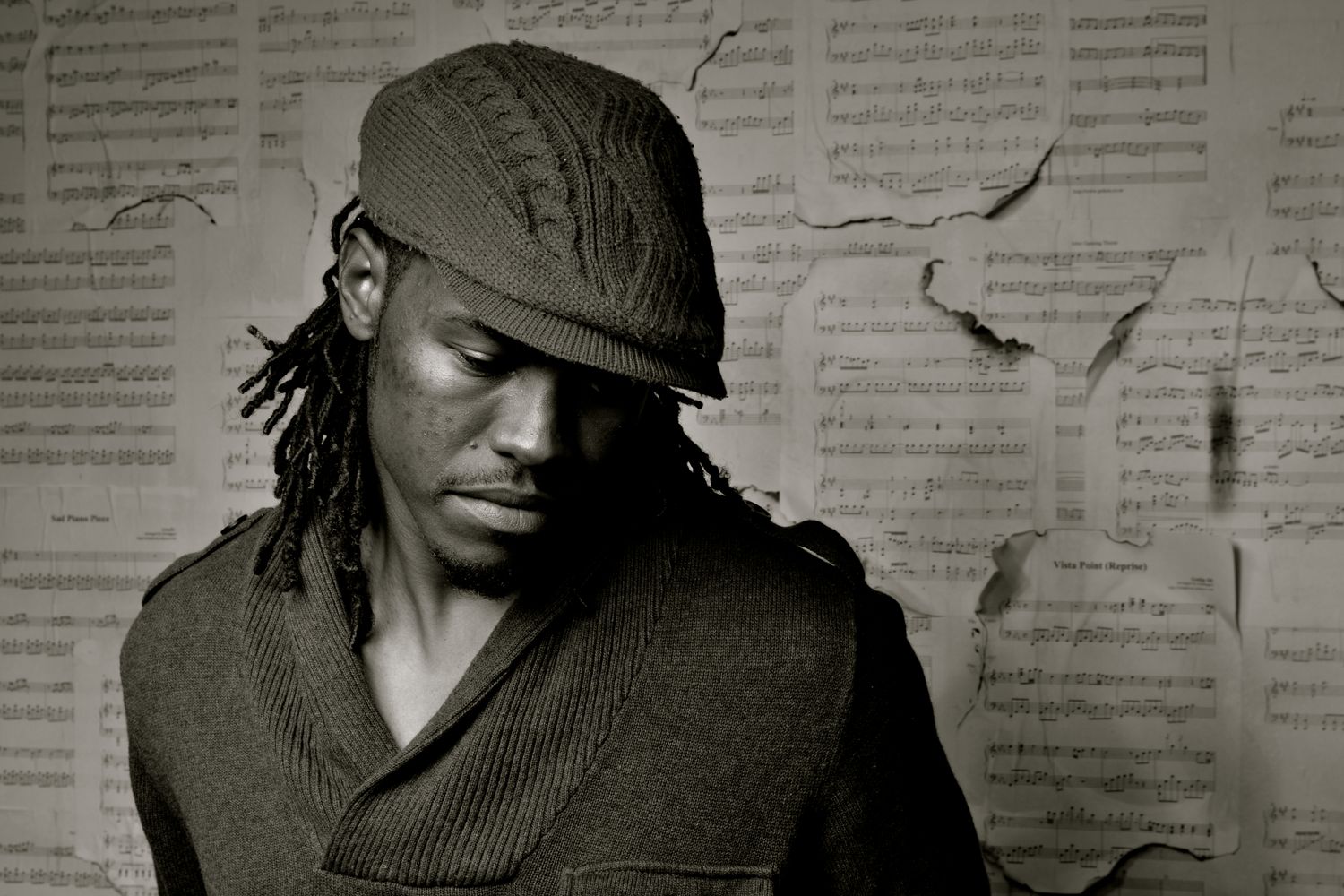 A man with dreadlocks and a knit cap poses thoughtfully against a wall covered in sheet music.