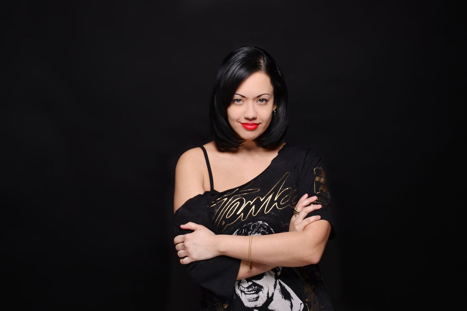 Woman with short black hair and red lips poses confidently in a black top against a dark background.