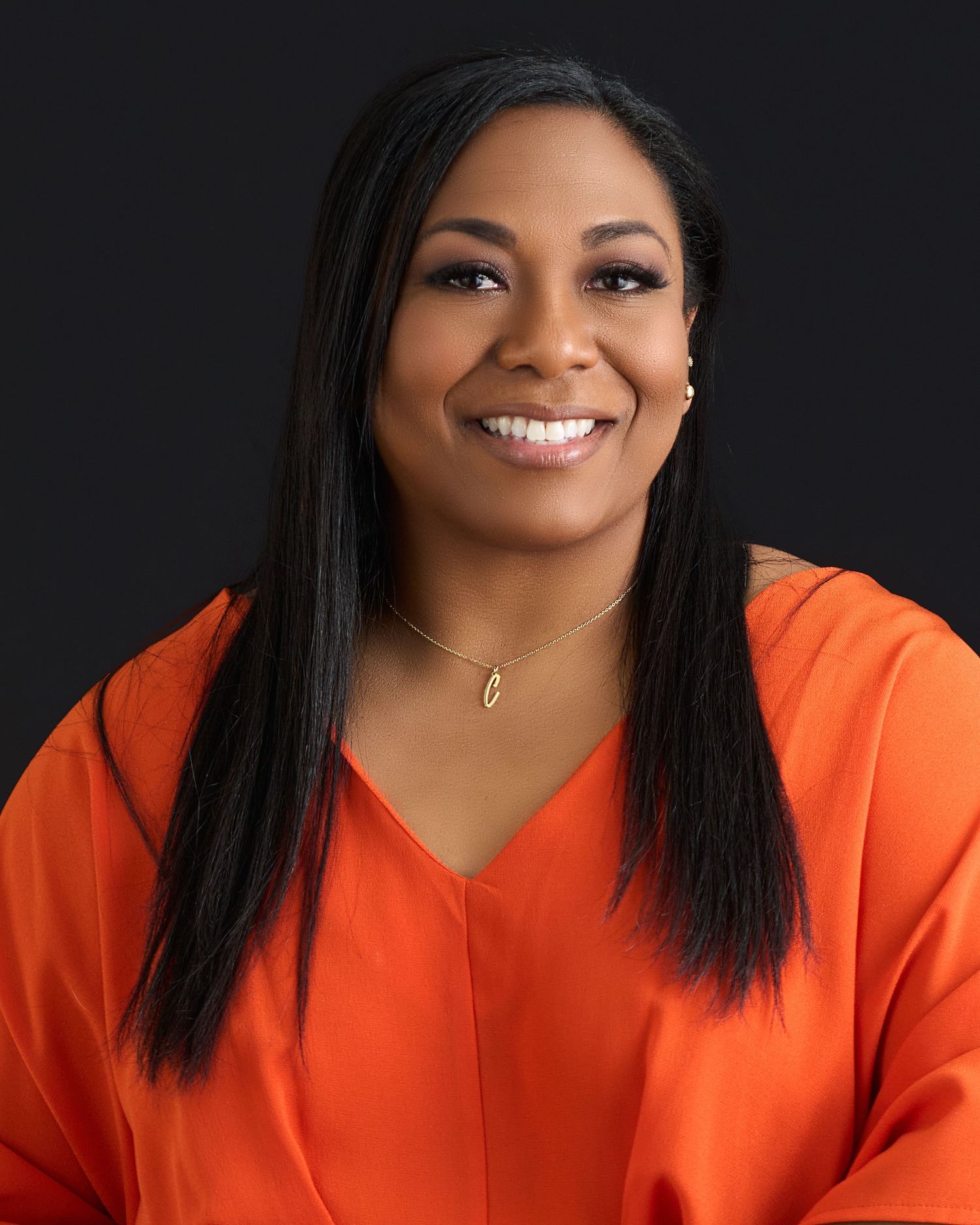 A smiling woman in an orange top with straight black hair and a gold necklace poses against a dark background.