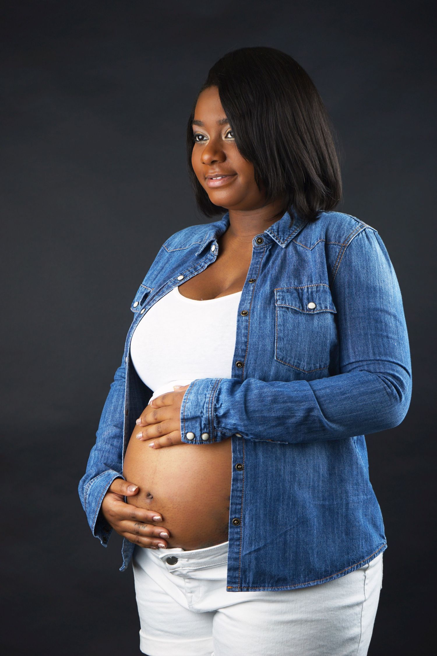 Pregnant Black woman in denim shirt holding baby bump, maternity portrait on dark background.