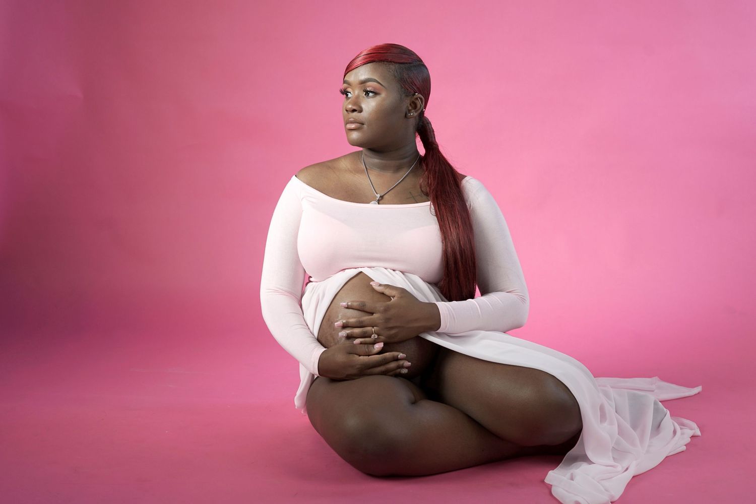 Pregnant woman in white dress sitting cross-legged against pink background.
