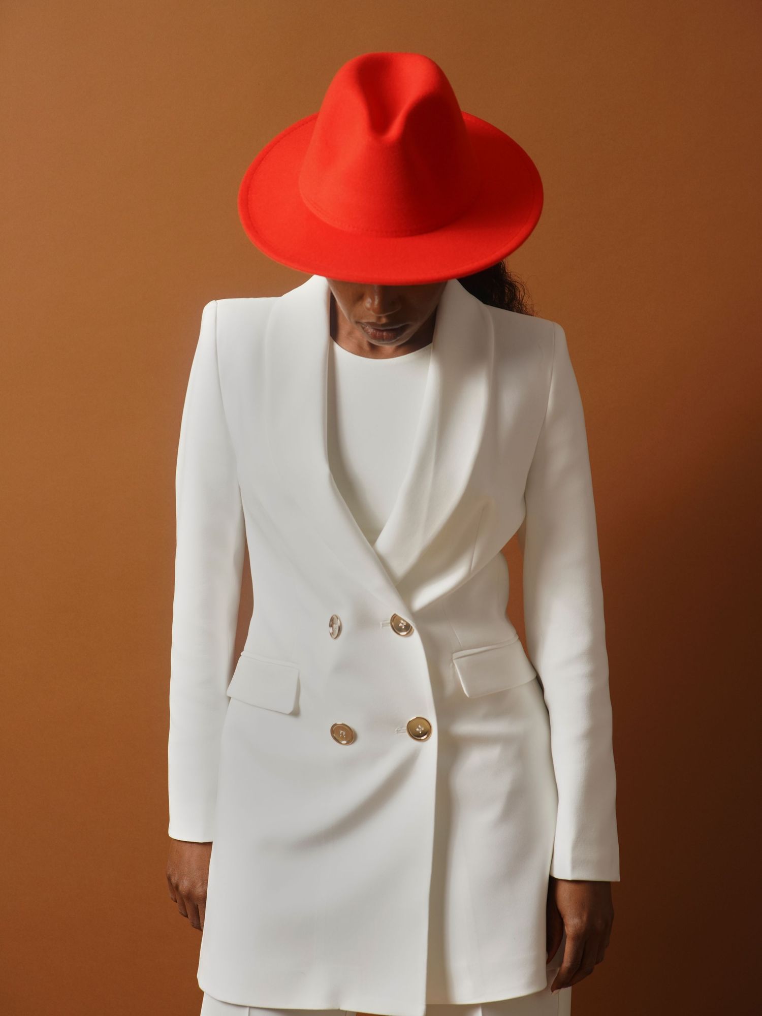 A stylish person in a white double-breasted blazer and bold red fedora hat poses against a brown background.