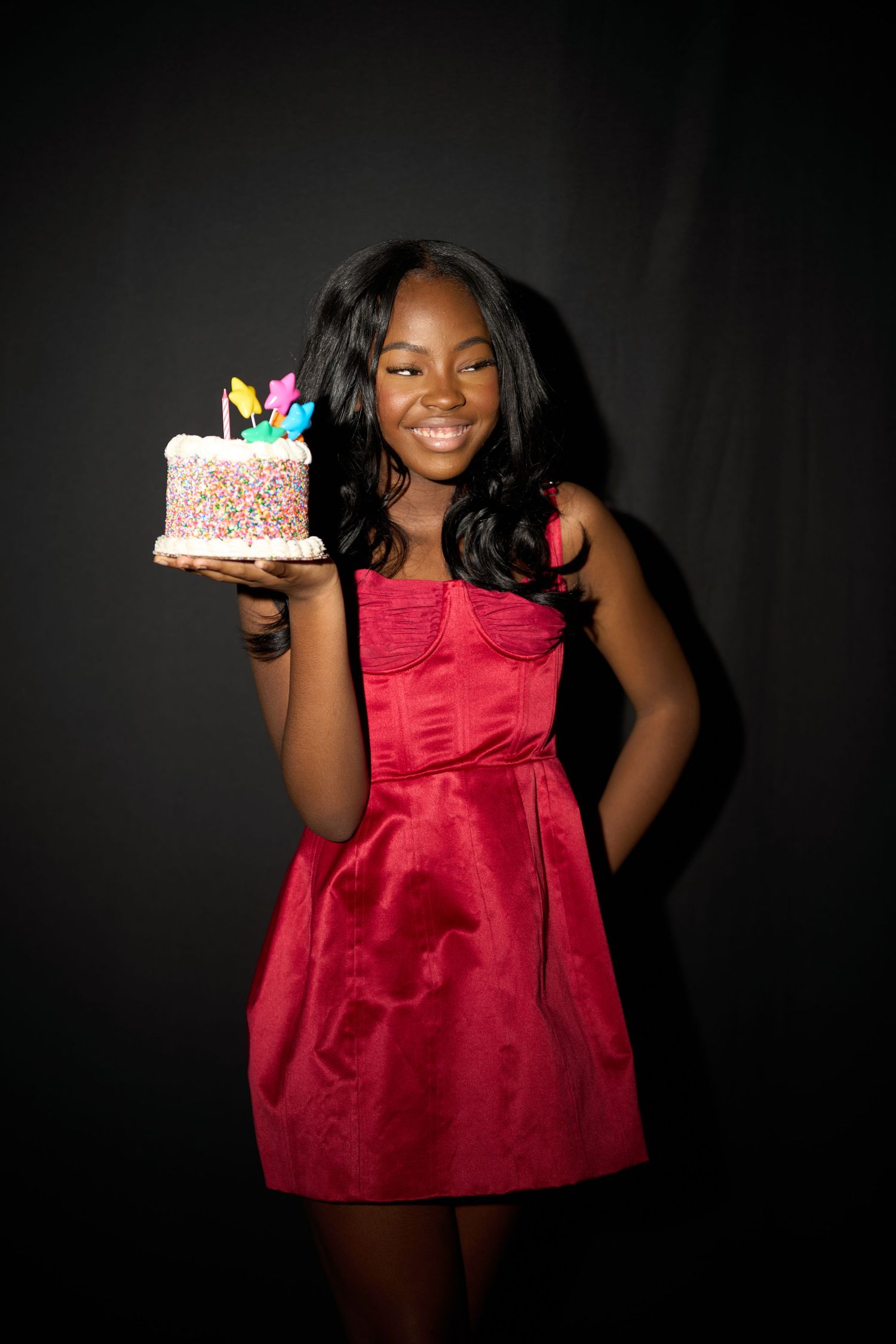 Smiling young girl in red dress holding colorful birthday cake with candles against dark background.