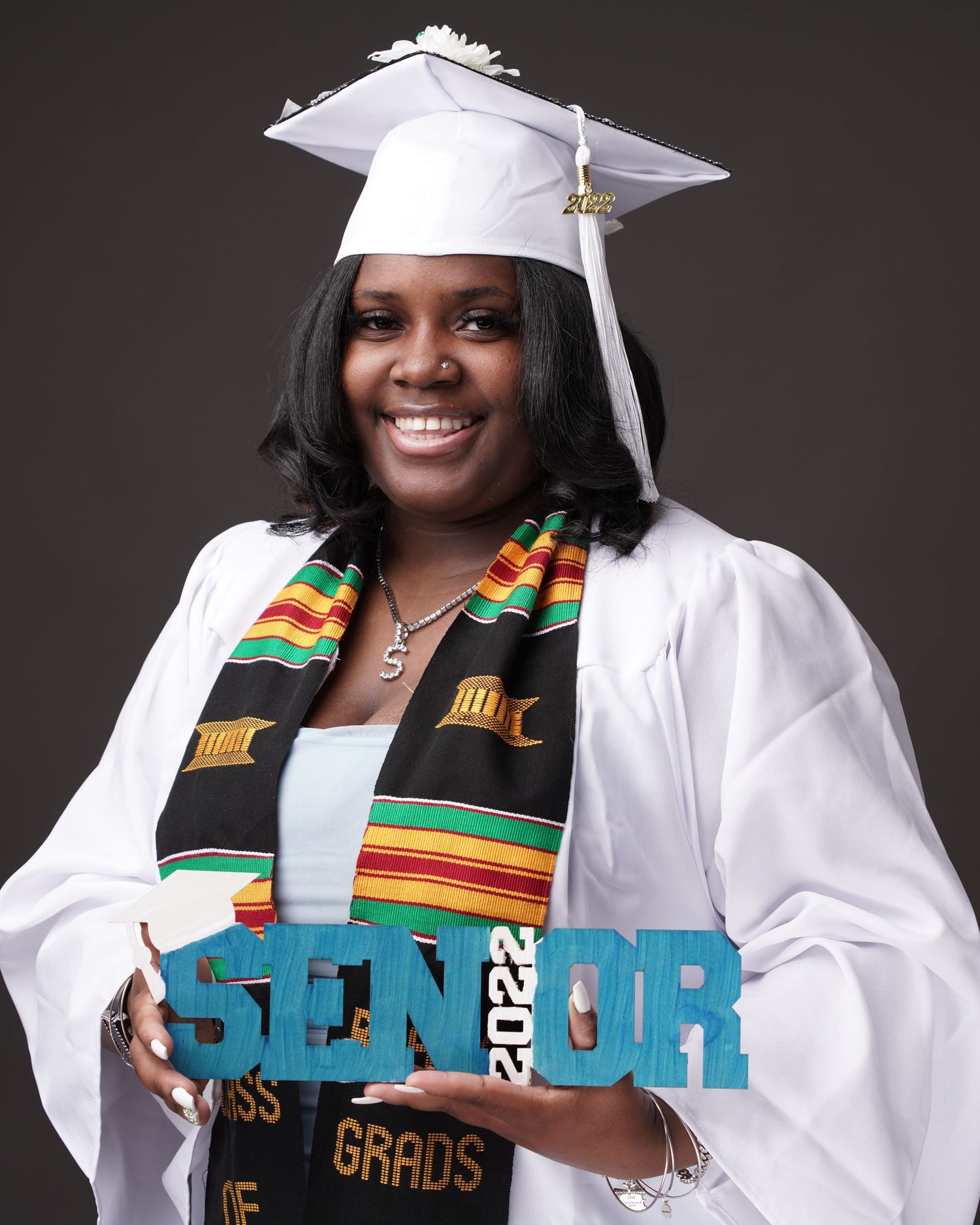 A smiling graduate in white cap and gown with a kente cloth stole holds a Senior 2022 sign.