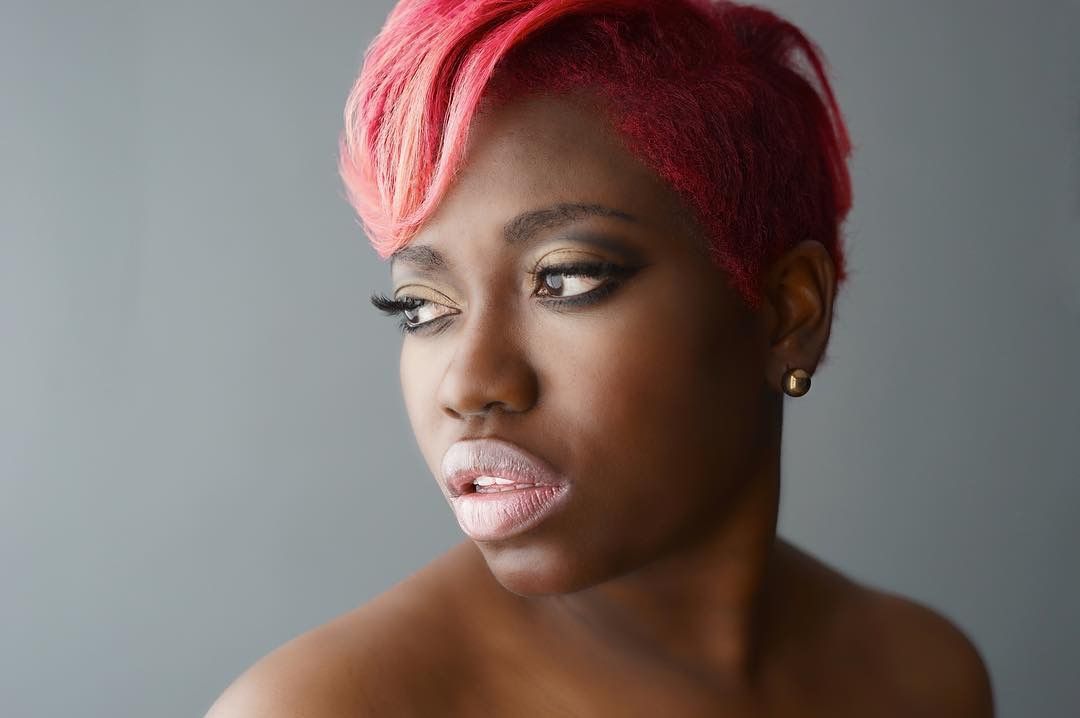 Young Black woman with short pink hair and bold makeup, artistic close-up portrait on gray background.