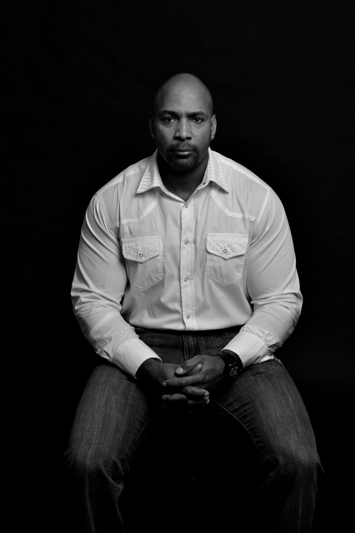 Serious bald Black man in white button-up shirt sitting with clasped hands in black and white portrait.