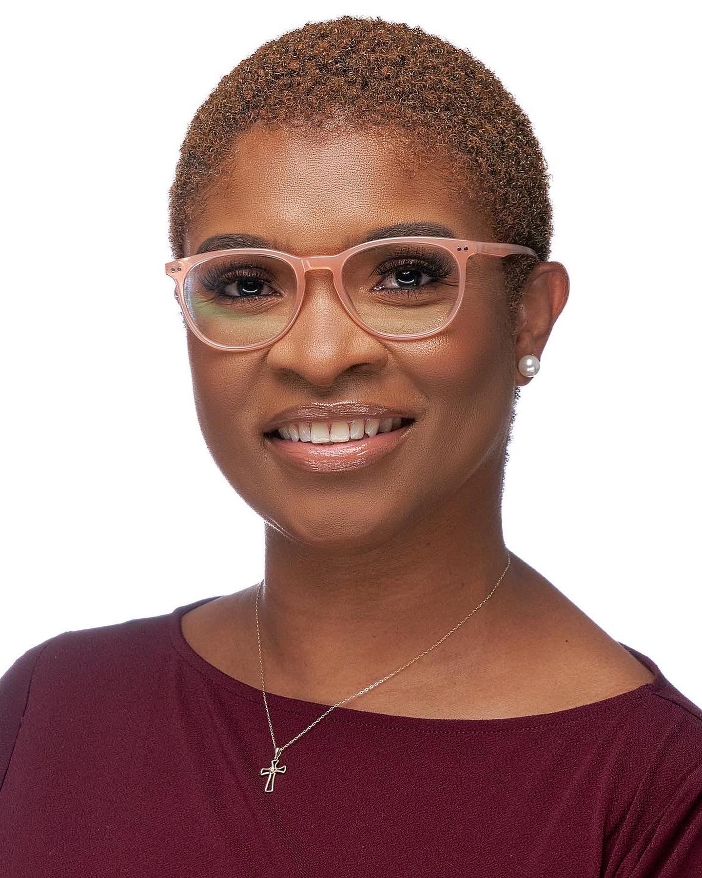 Woman with short natural hair wearing pink glasses and cross necklace smiling.