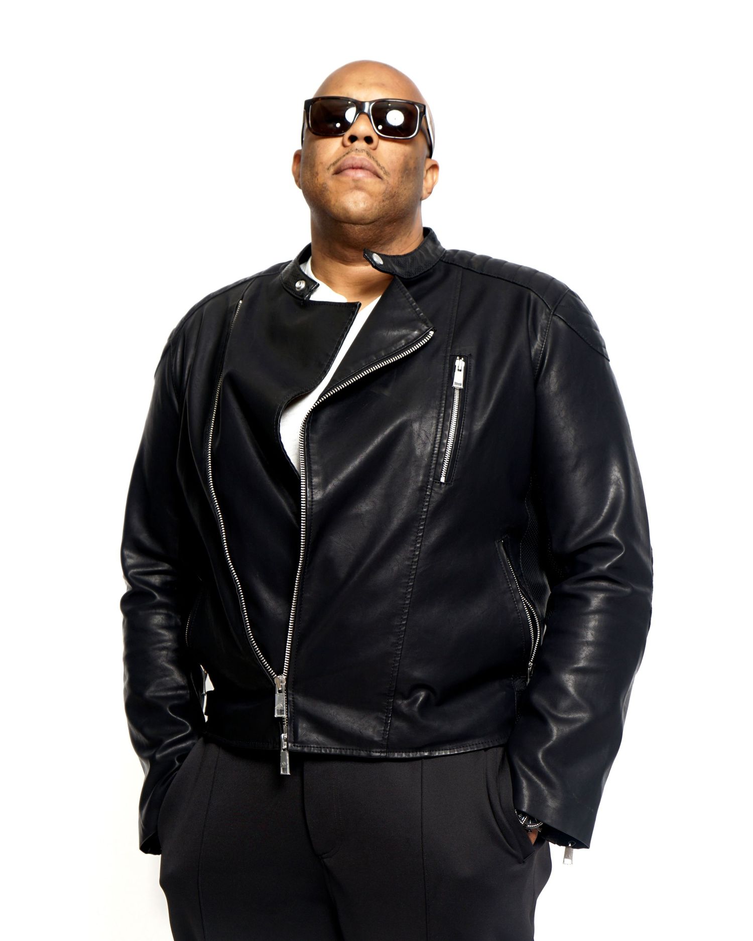 Man in black leather biker jacket and sunglasses posing confidently against white background.