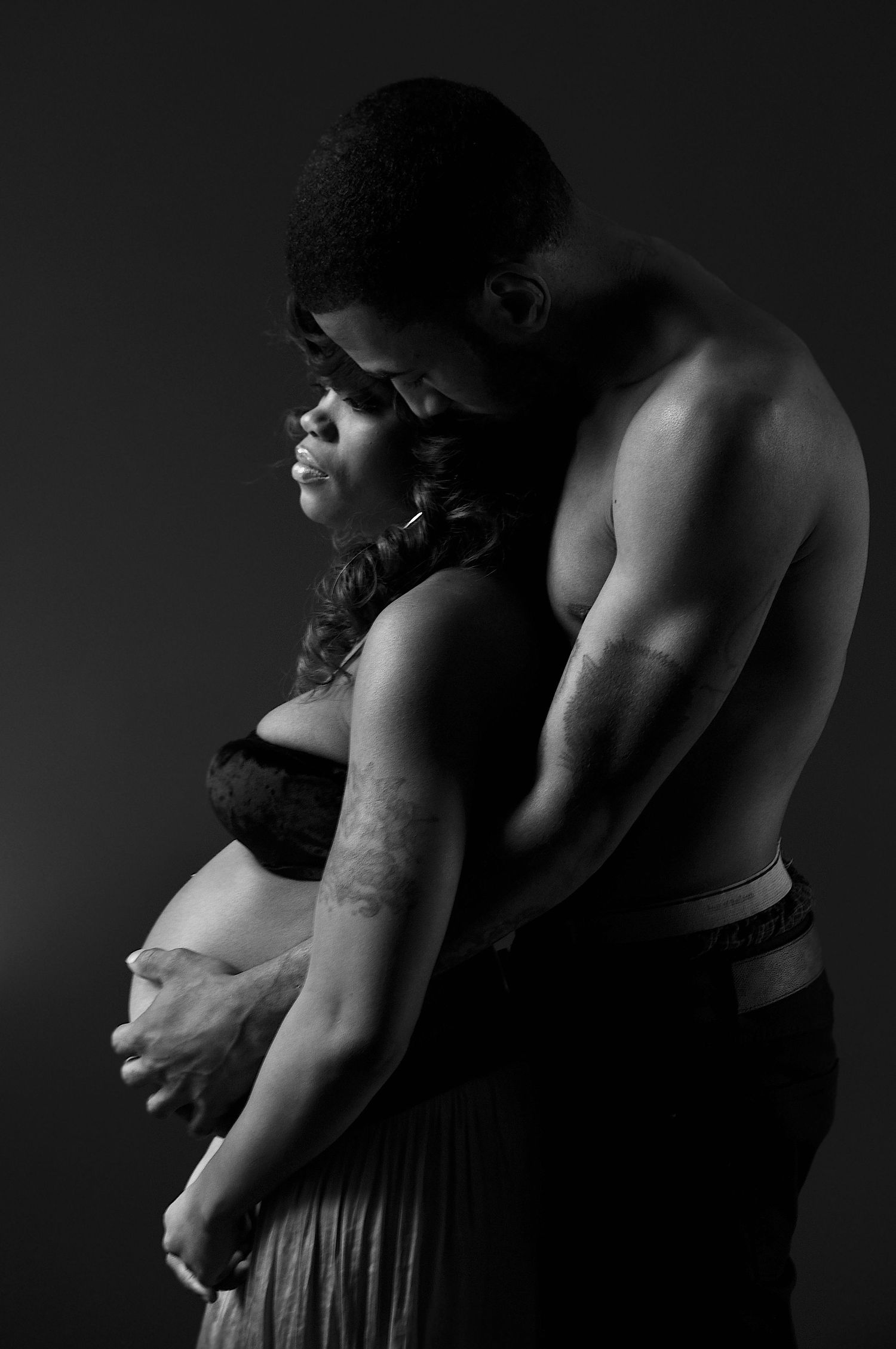Black and white maternity photo of a couple embracing tenderly in a dark studio setting.