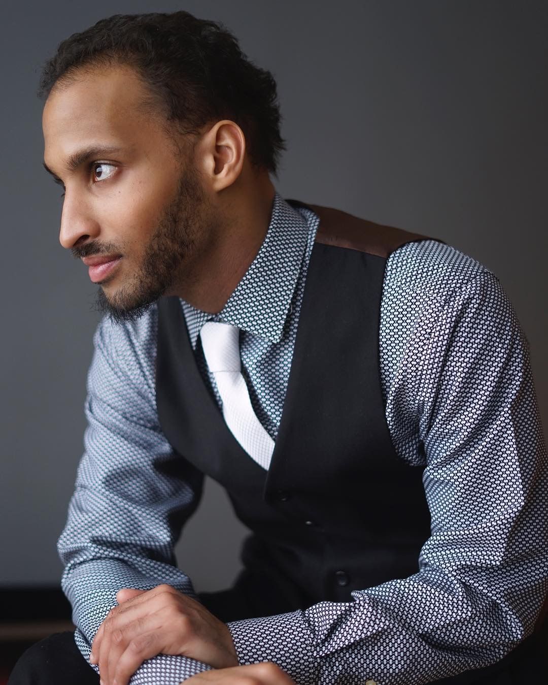 Young man in black vest and patterned shirt looking sideways, moody studio portrait on gray background.