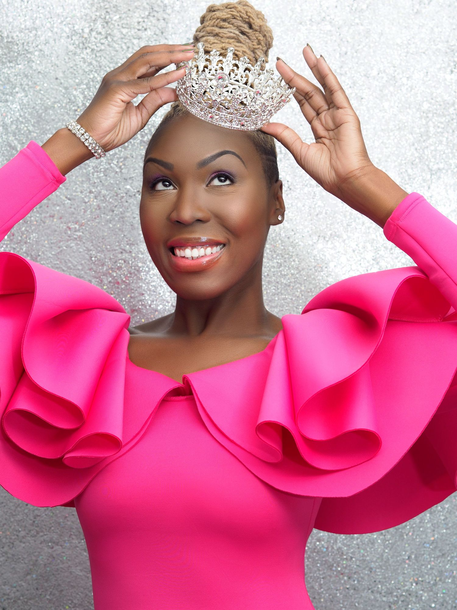 A joyful Black woman in a bright pink ruffled dress places a sparkling tiara on her head, smiling confidently.