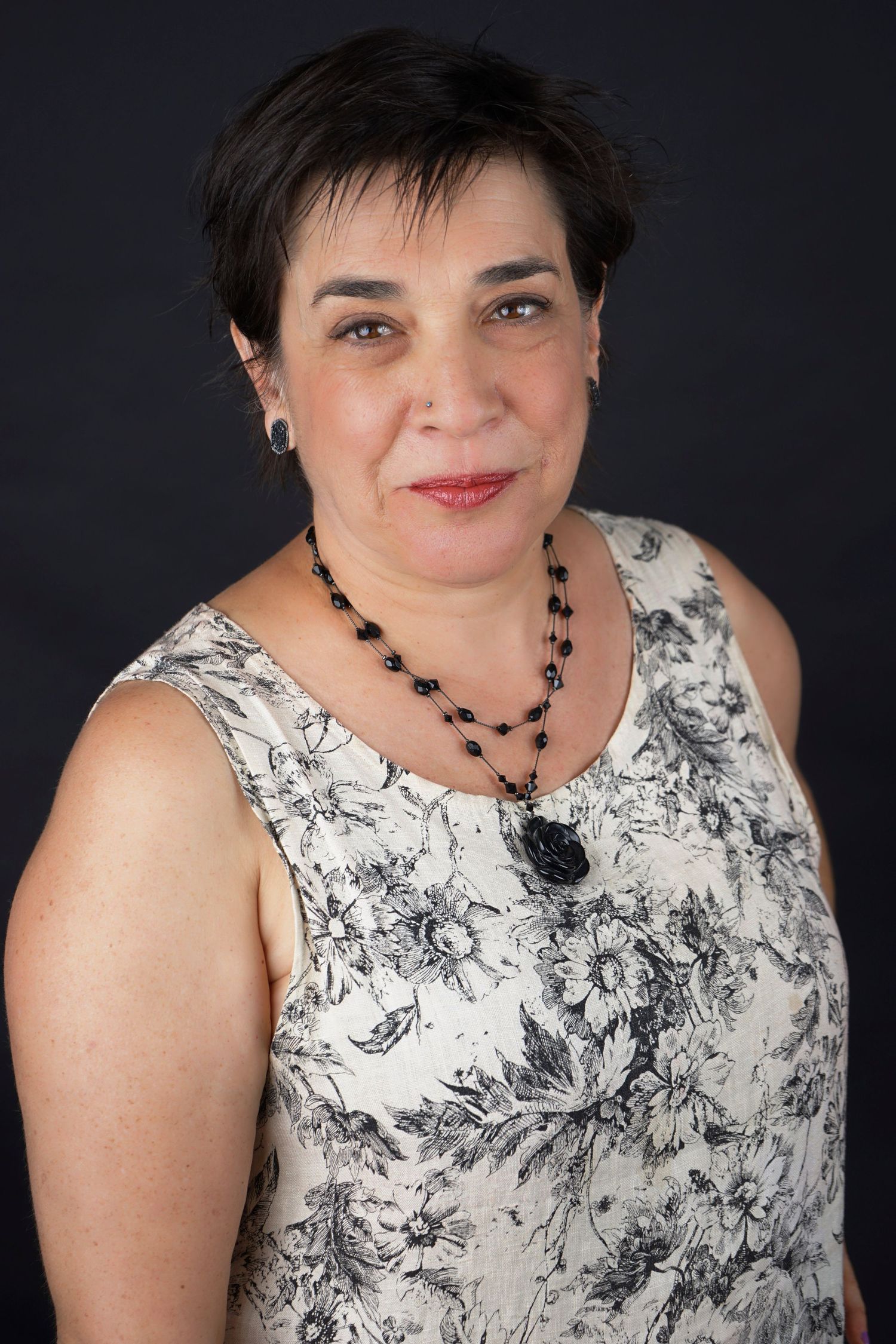 Middle-aged woman with short dark hair wearing floral dress and black beaded necklace on dark background.
