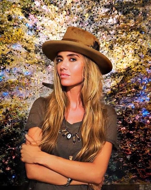 Woman with long blonde hair wearing a wide-brim hat and necklace posing before a floral backdrop.