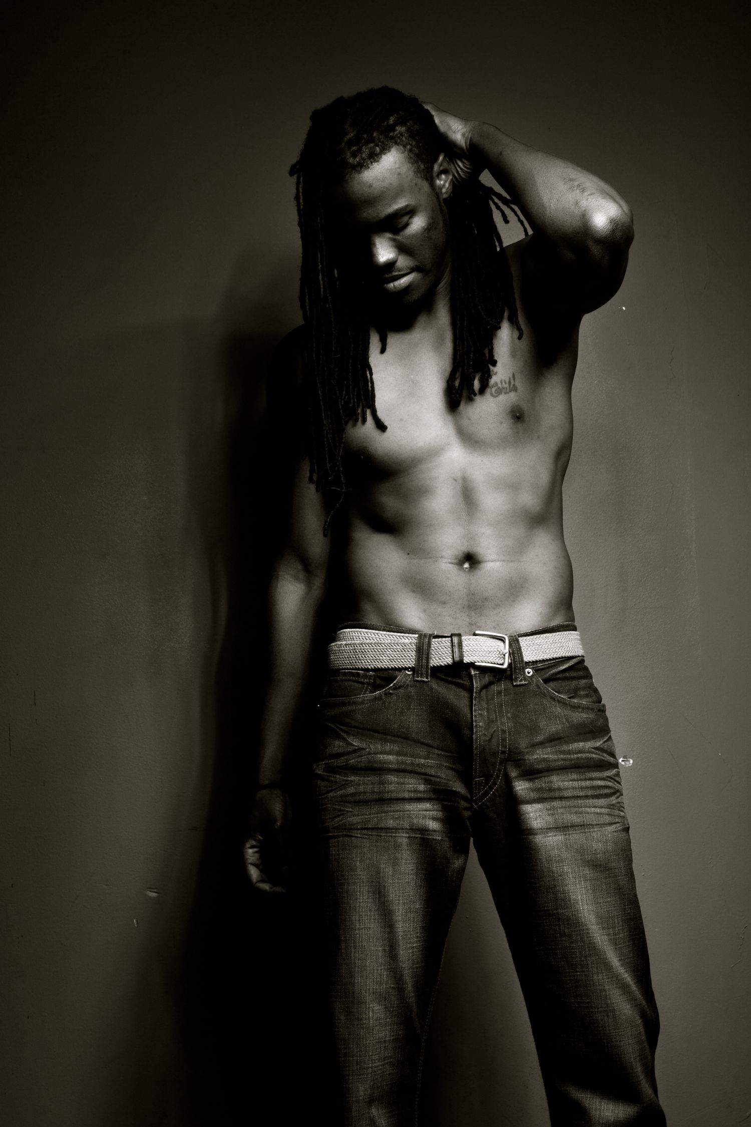 A shirtless man with long dreadlocks poses in jeans against a dark wall in a black-and-white photo.