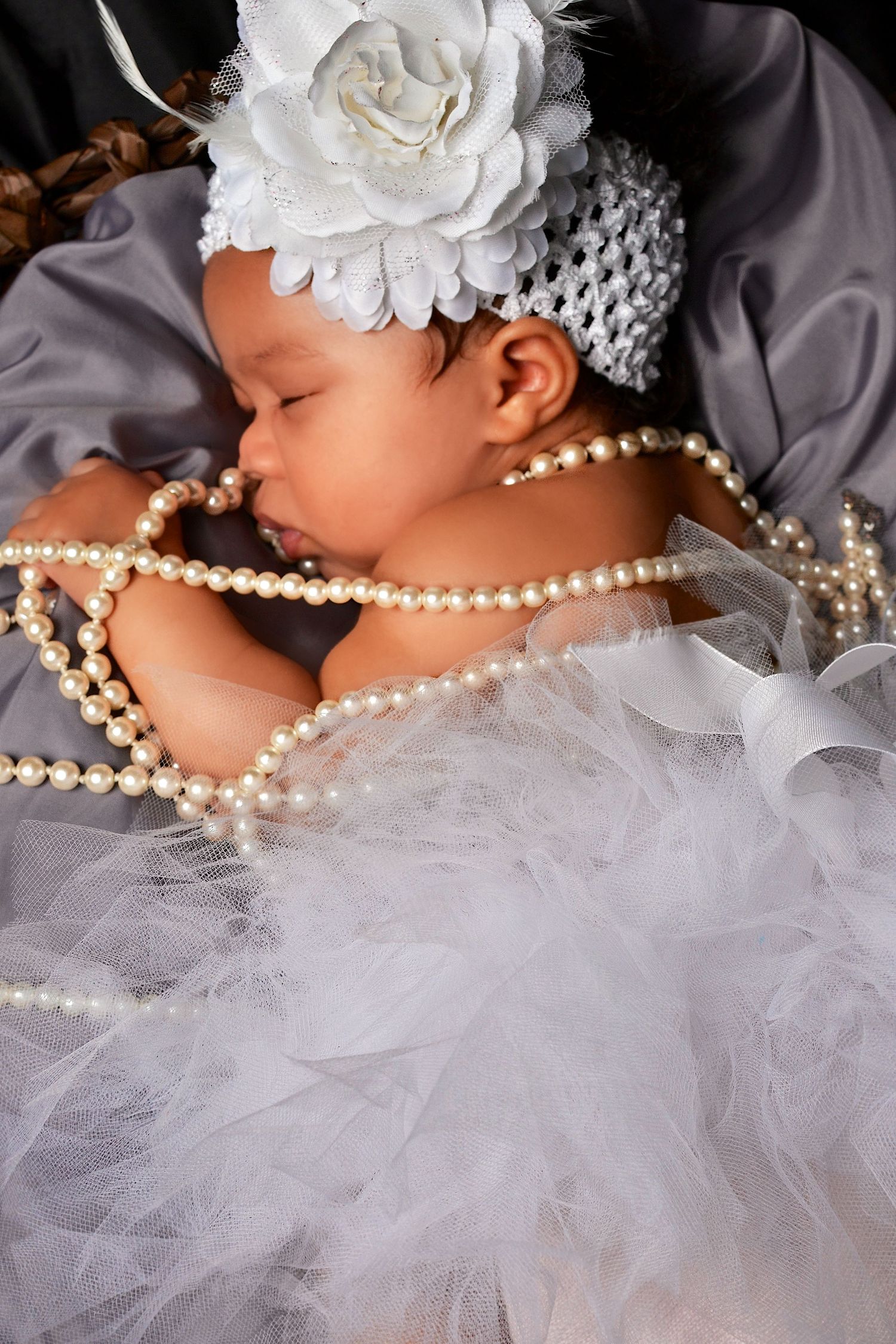 Sleeping Black newborn baby girl dressed in white tulle with pearl necklace and floral headband.