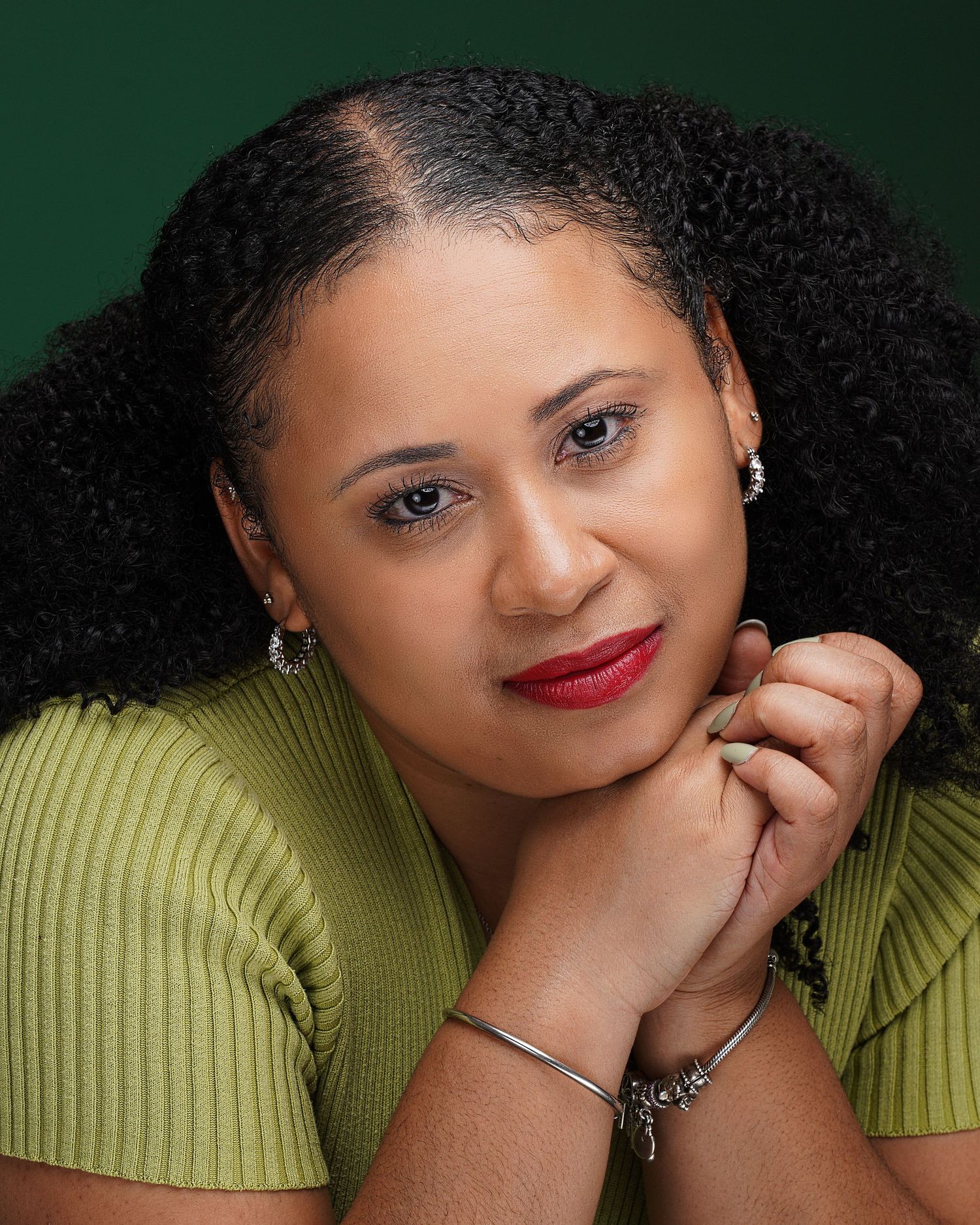 A woman with natural curly hair poses gracefully in a green ribbed top with red lips and silver jewelry.